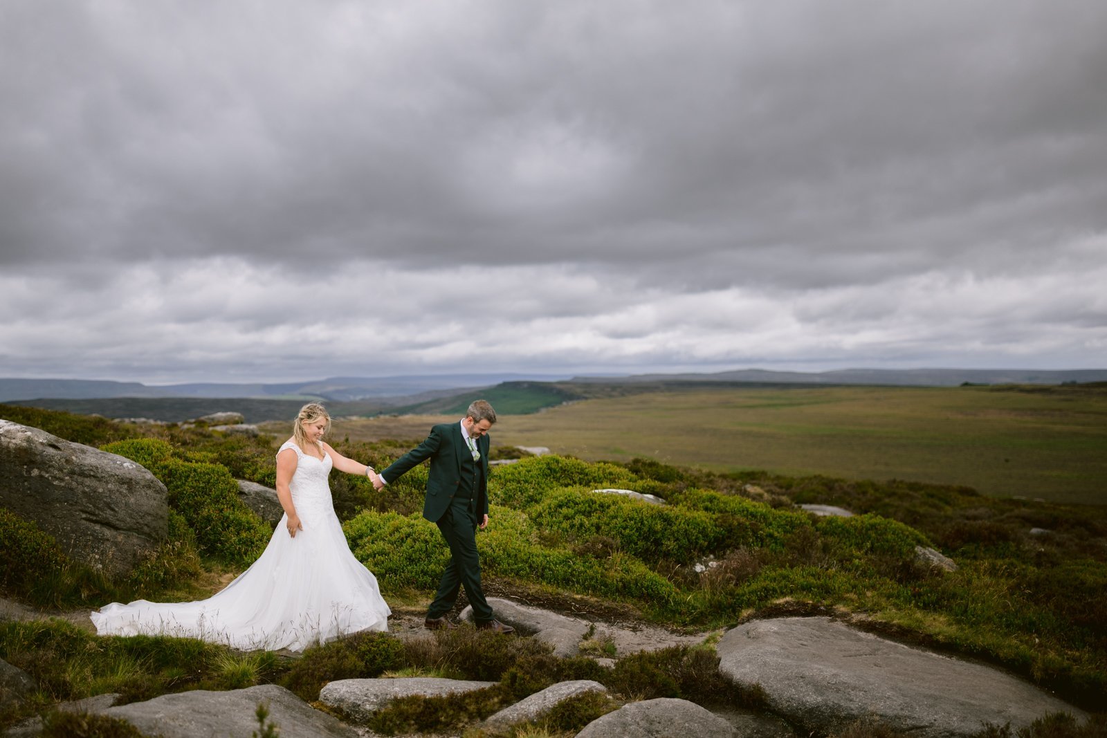 A bride and groom walk hand in hand across a rocky, grassy landscape under a cloudy sky.
