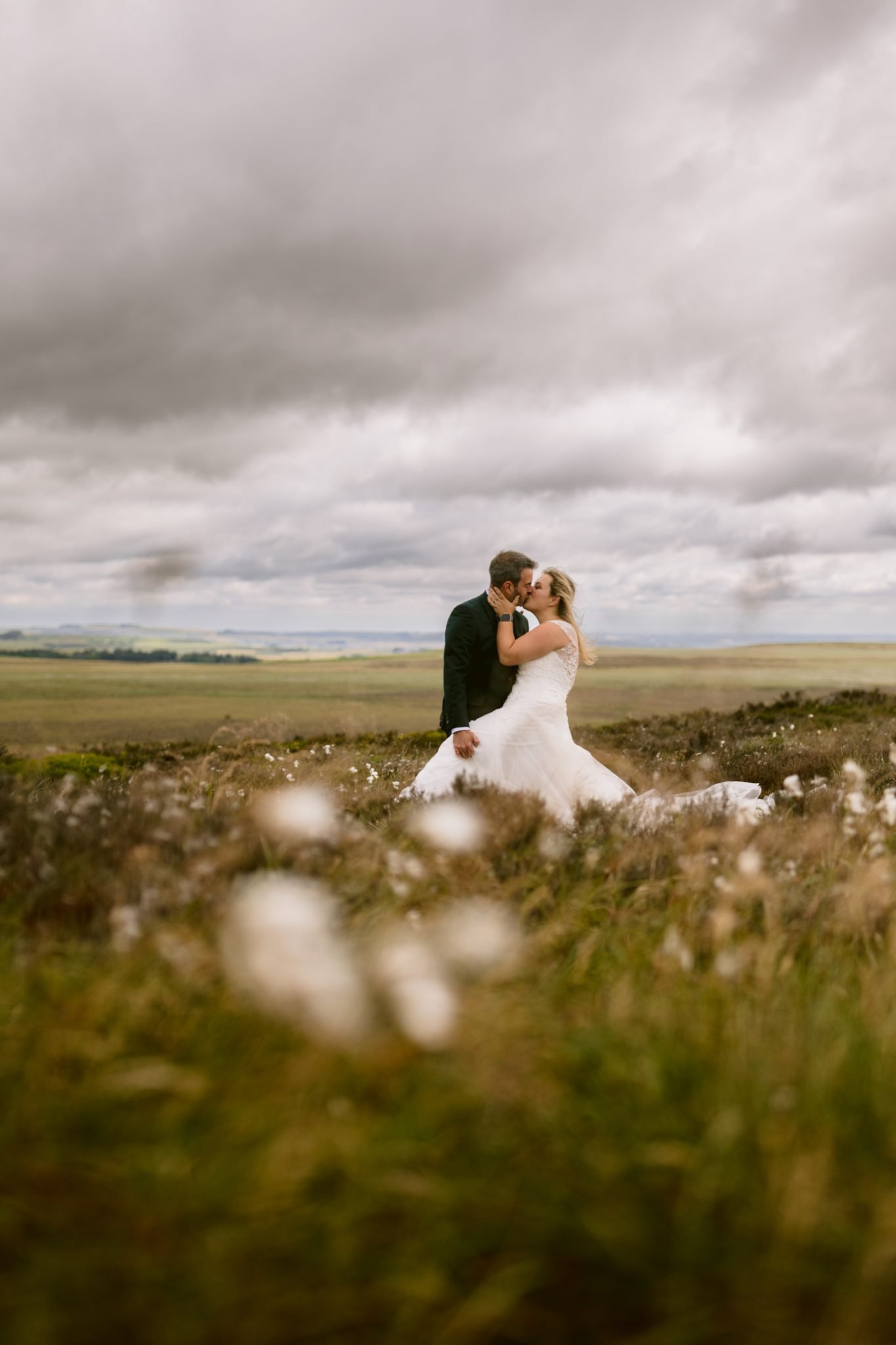 A bride and groom stand in a grassy field, embracing and kissing under a cloudy sky.