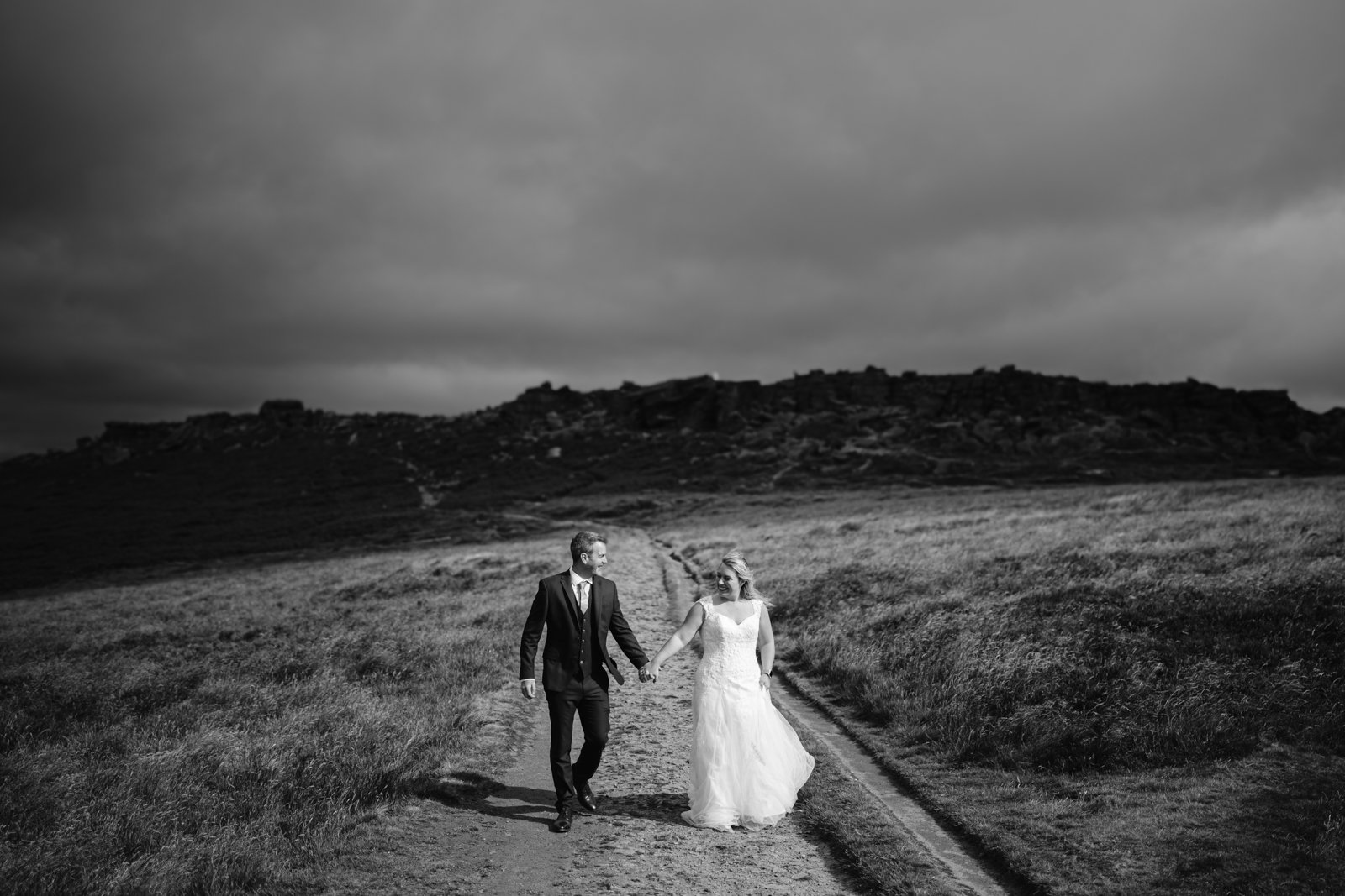 A bride and groom walk hand in hand on a dirt path through a grassy field under a cloudy sky, with rocky hills in the background.
