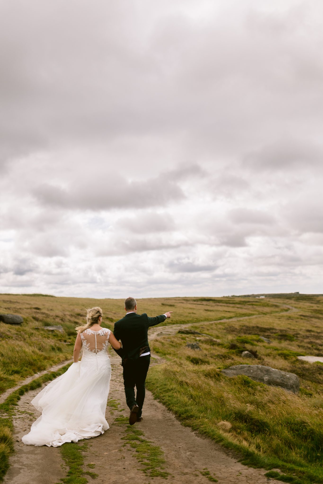 A bride and groom walk down a dirt path through grassy fields under a cloudy sky, with the groom pointing ahead.
