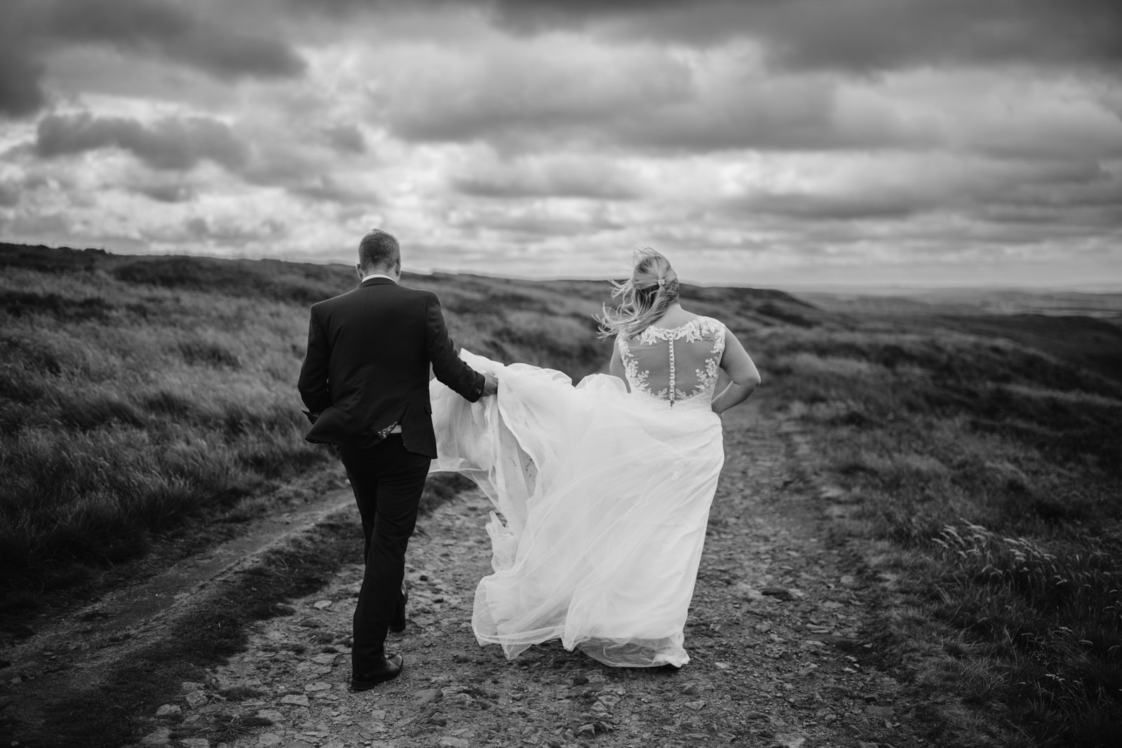 A couple in wedding attire walks along a rocky path in a rural landscape under a cloudy sky.