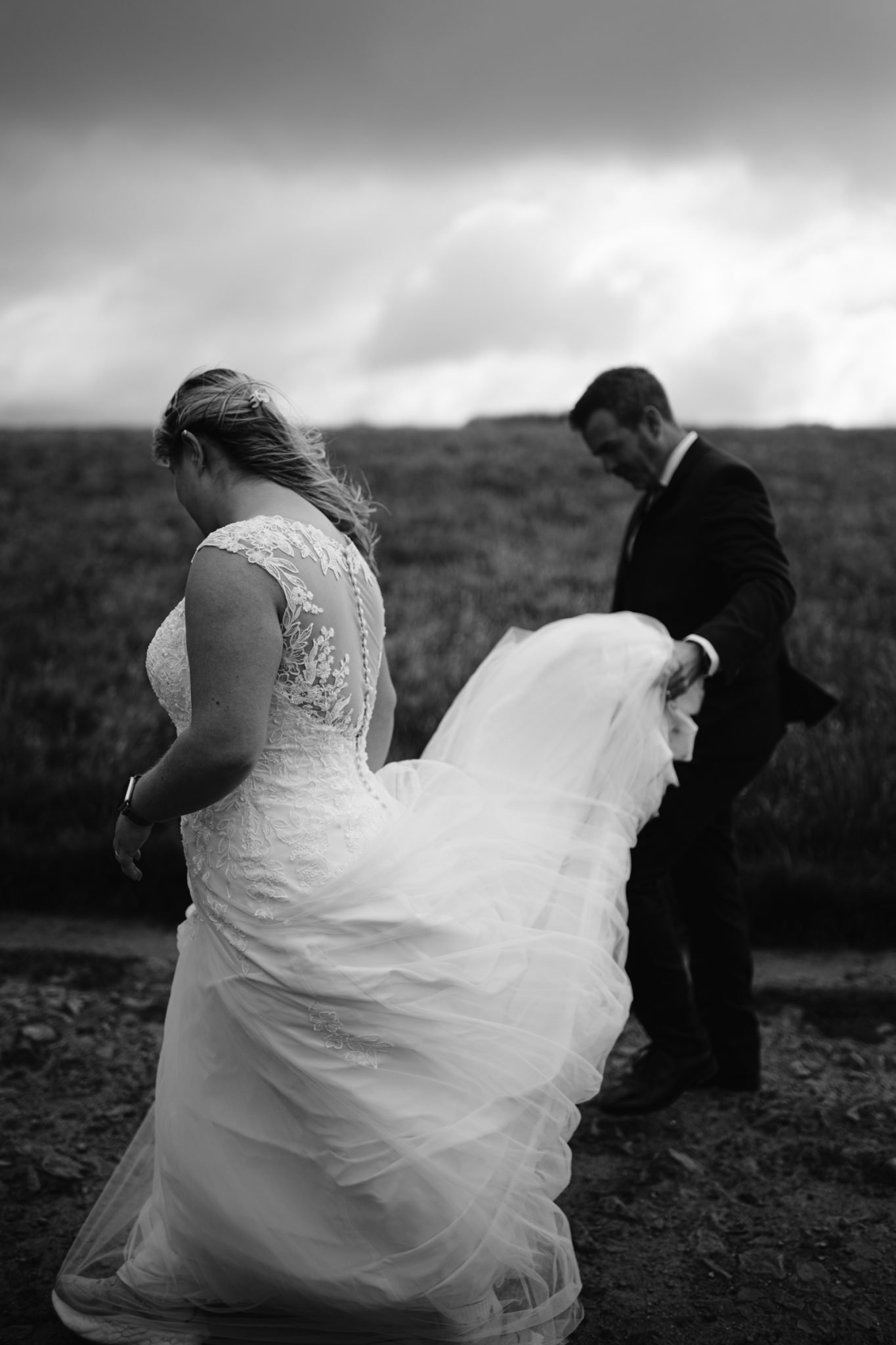 A bride in a lace wedding dress walks outdoors as a groom in a suit holds her train. The scene is in black and white with cloudy skies in the background.