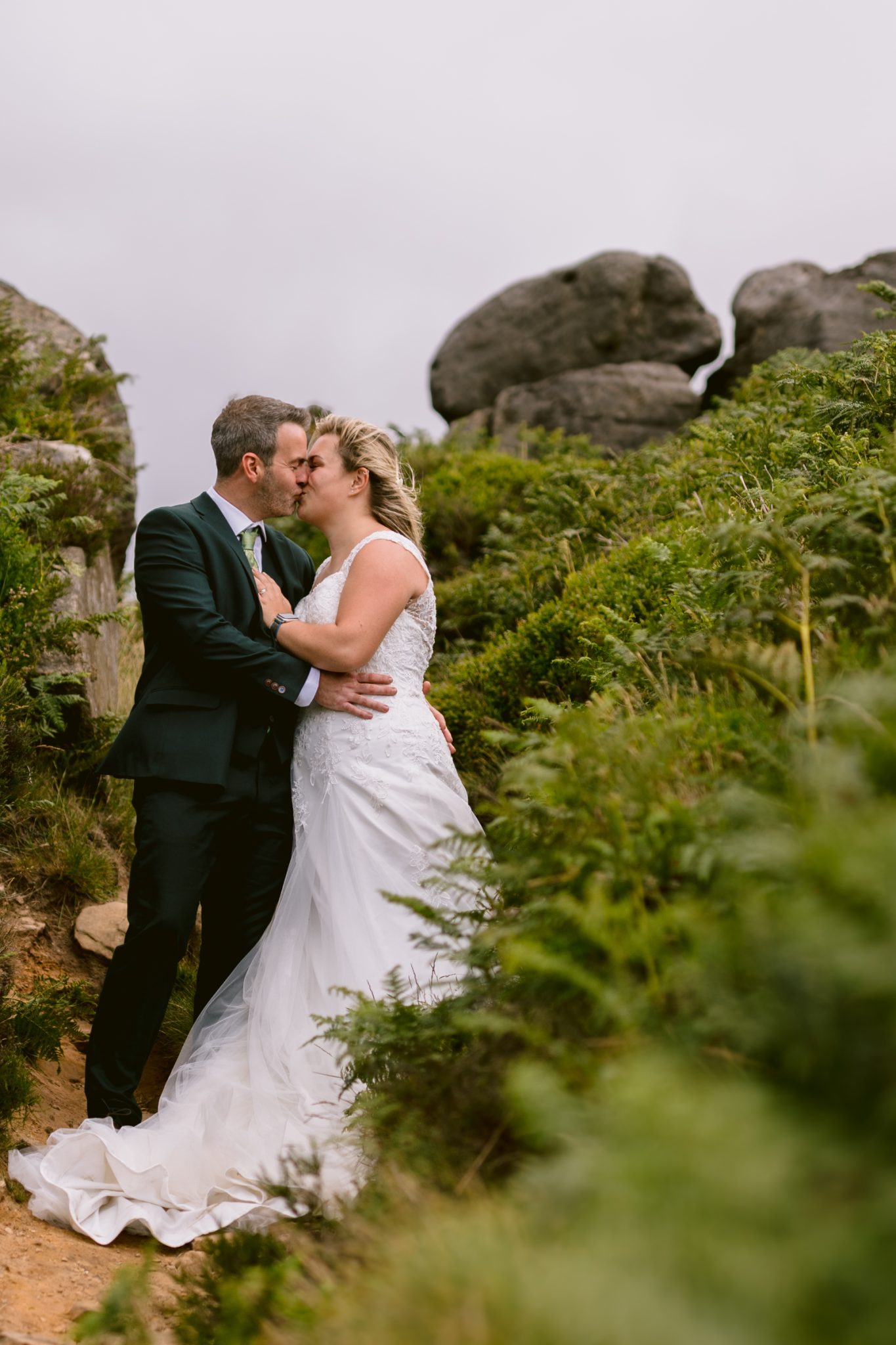 A bride and groom in wedding attire share a kiss outdoors, standing on a dirt path surrounded by greenery and large rocks.