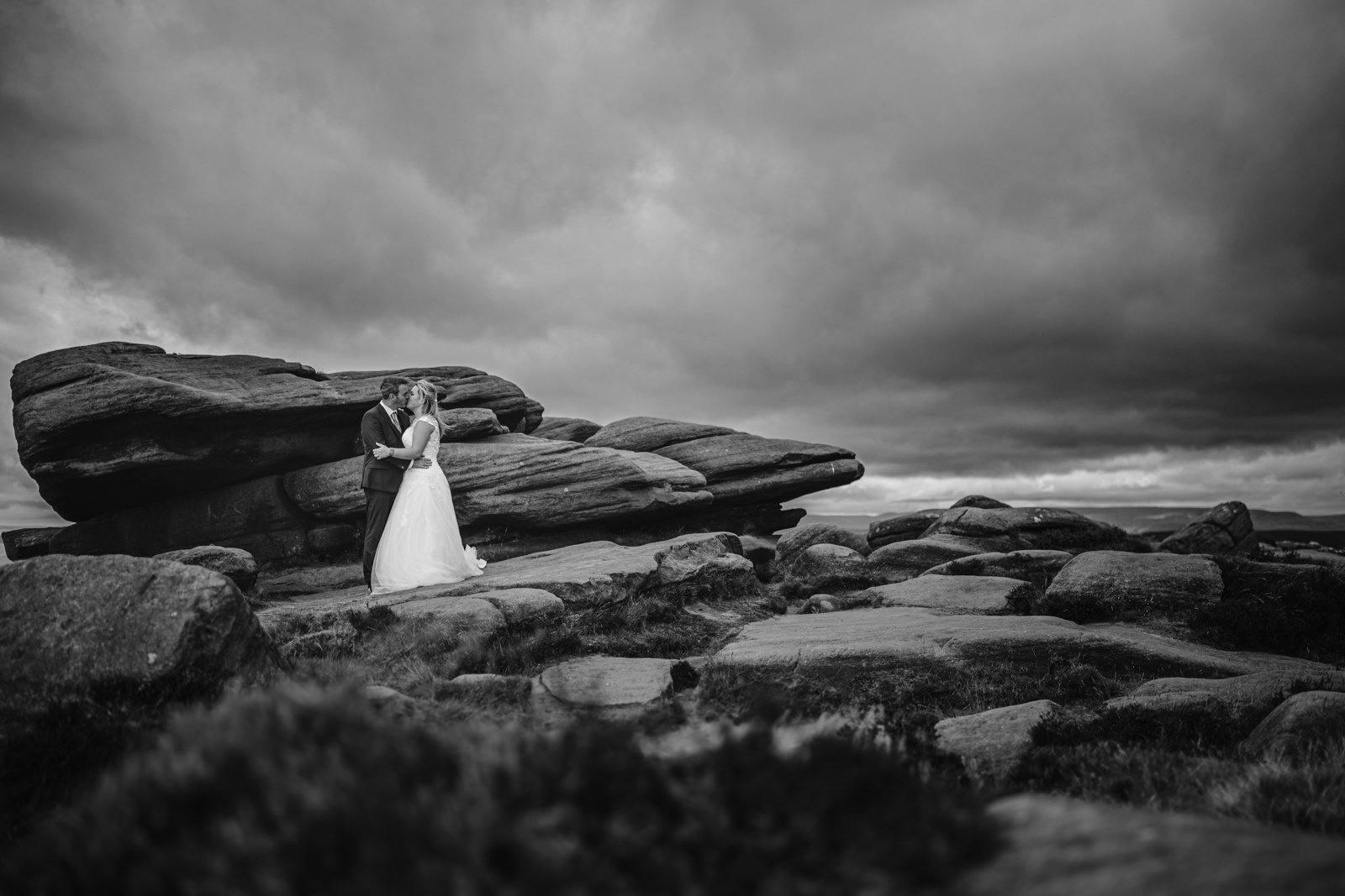 A bride and groom stand embracing on rocky terrain under a cloudy sky, surrounded by large stone formations.