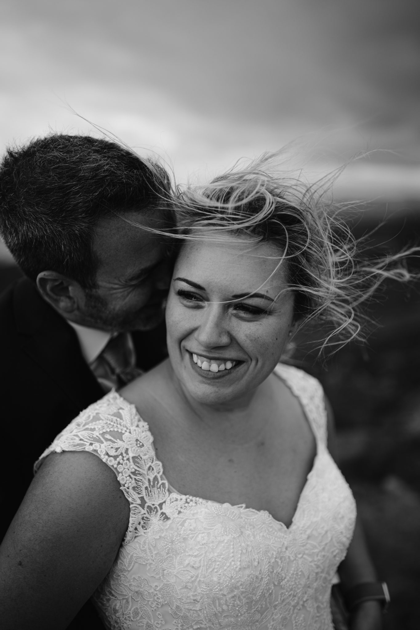 A smiling bride in a lace dress stands in front of a groom, who is leaning in close. The wind blows her hair across her face. The photo is black and white.