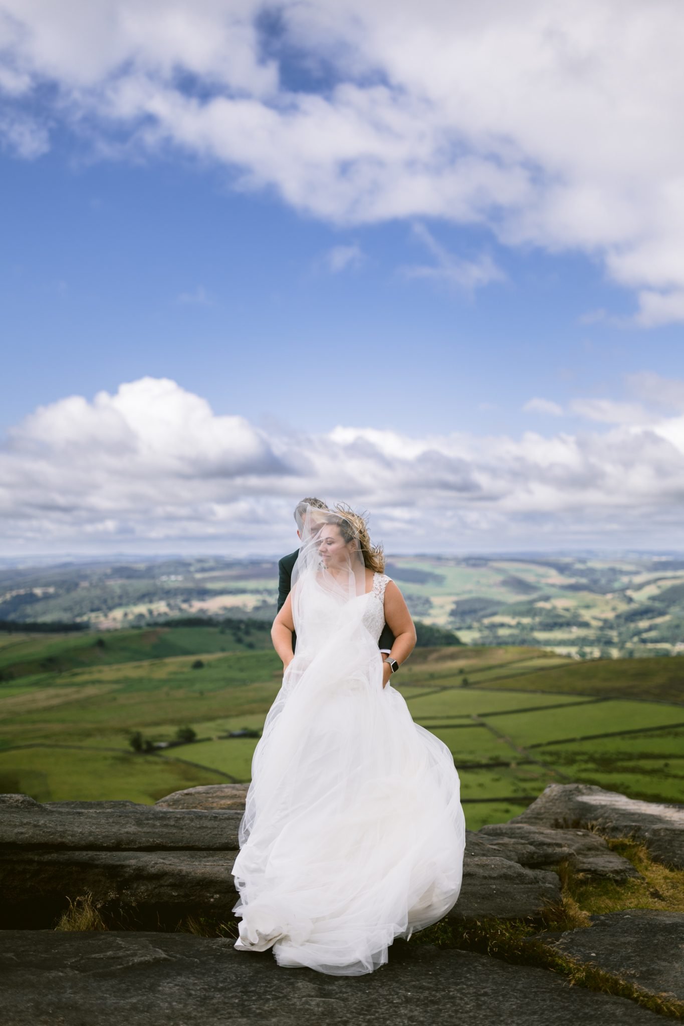 A bride in a flowing white dress stands on rocky ground with a groom behind her, set against a backdrop of rolling green hills and a partly cloudy sky.