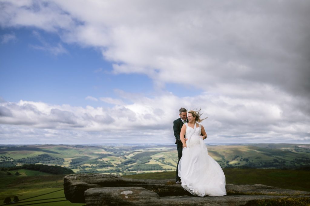 A bride and groom stand on a rocky ledge overlooking rolling green hills under a cloudy sky with patches of blue.