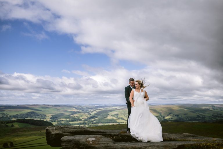A bride and groom stand on a rocky ledge overlooking rolling green hills under a cloudy sky with patches of blue.