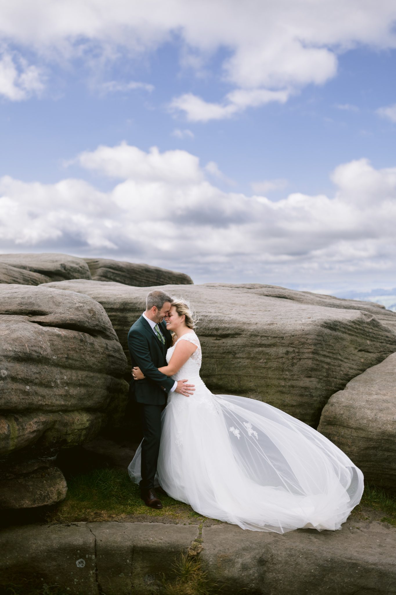 A bride and groom embrace and smile while standing on rocky terrain under a partly cloudy sky.