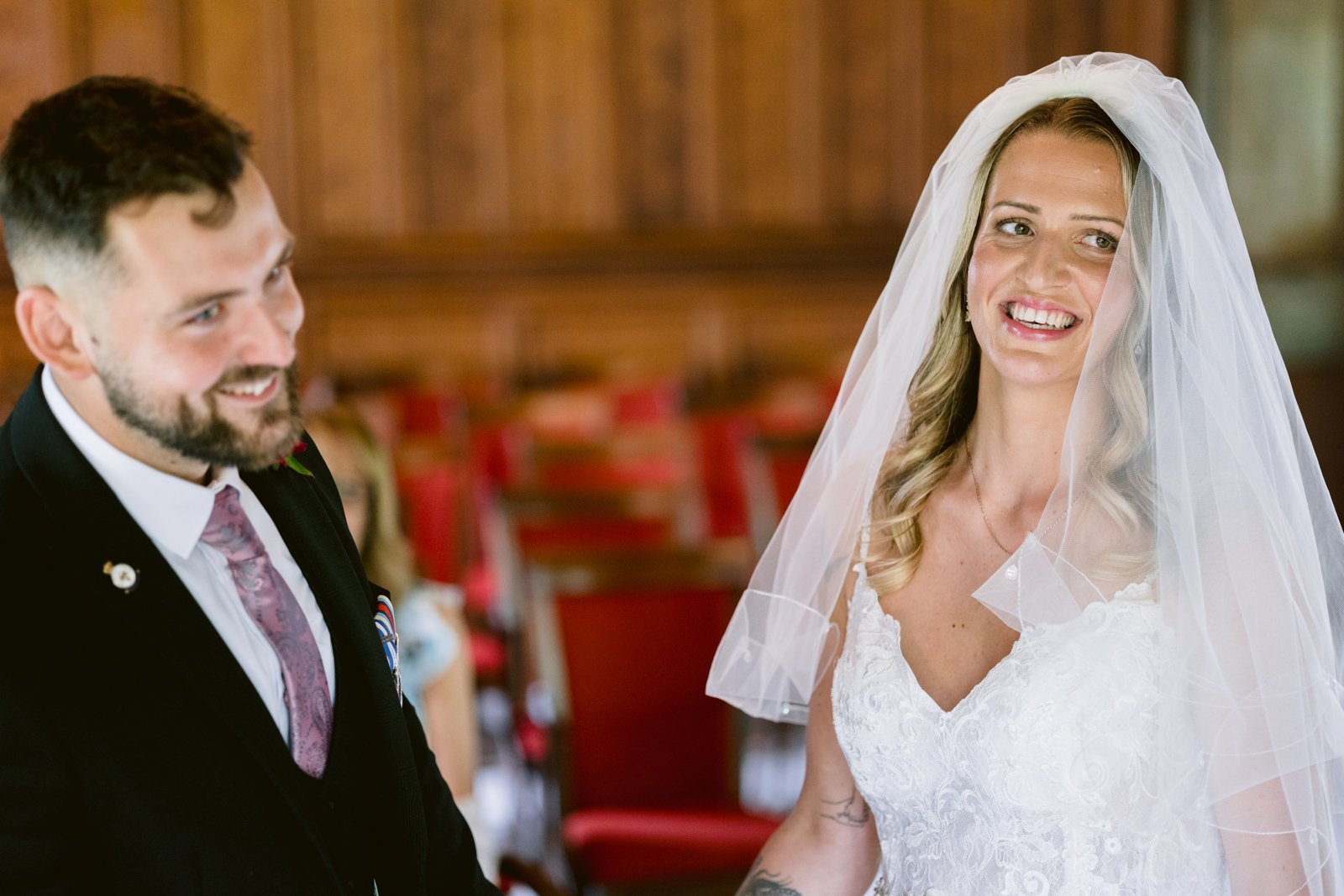 A bride in a white dress and veil stands smiling next to a groom in a suit and tie, with red chairs and wooden walls in the background.