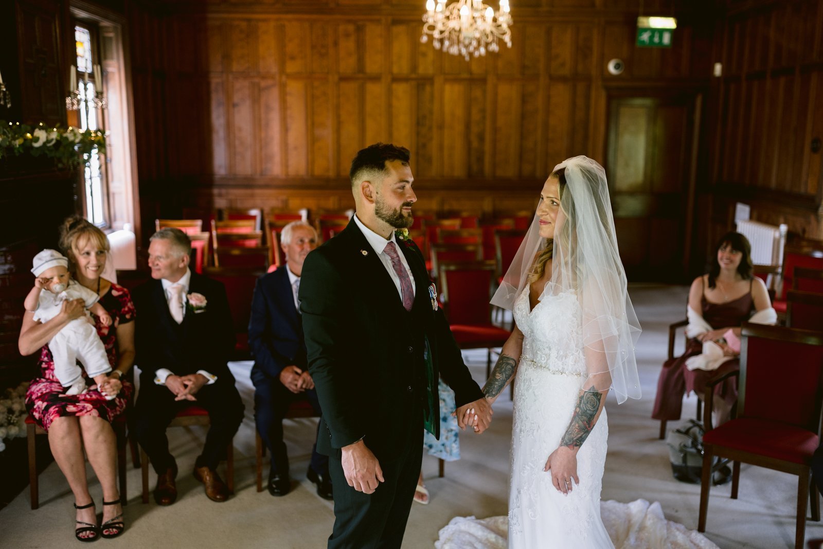 A bride and groom stand facing each other holding hands during a wedding ceremony in a wood-paneled room, with guests seated and watching.