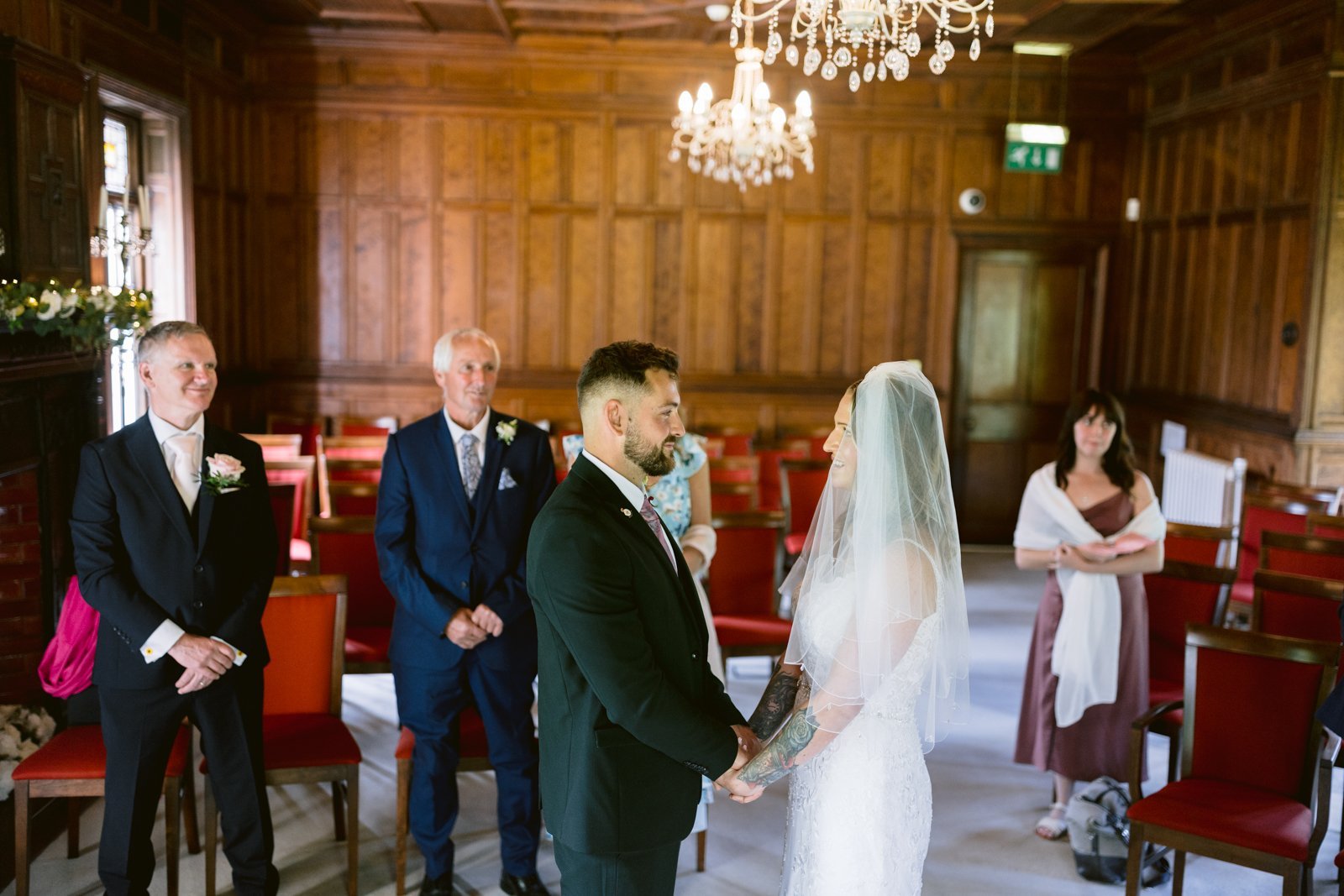 A bride and groom stand facing each other holding hands during a wedding ceremony in a wood-paneled room, with three guests watching.