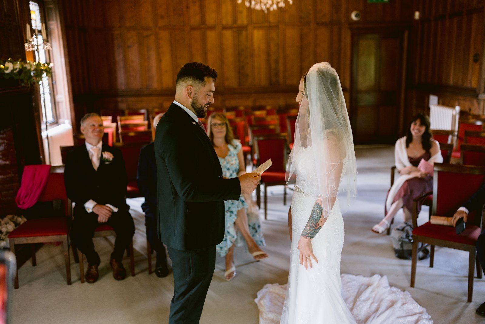 A groom reads vows to his bride during a small wedding ceremony in a wood-paneled room, with a few seated guests observing.