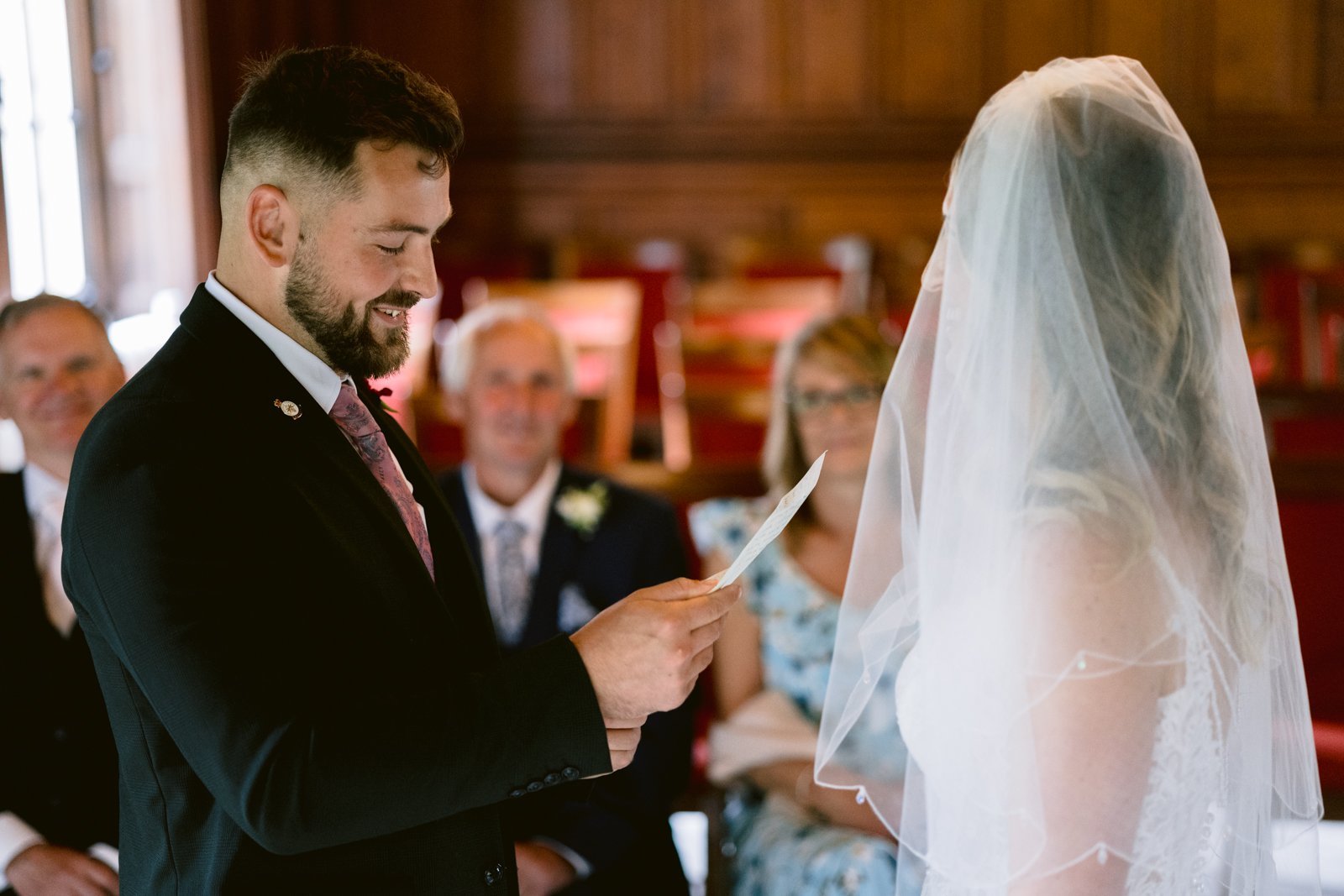 A groom in a suit reads from a piece of paper to a bride in a veil during a wedding ceremony, with seated guests watching in the background.