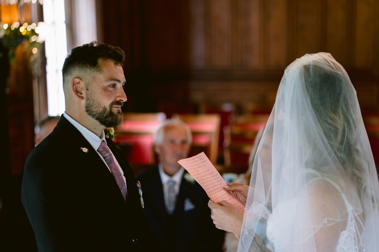 A bride in a veil reads from a piece of paper to a groom in a suit during a wedding ceremony in a wood-paneled room. An older man sits in the background.