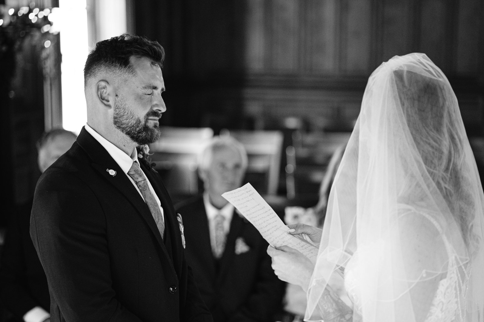 A bride reads vows from a piece of paper to the groom during a wedding ceremony in a formal indoor setting.