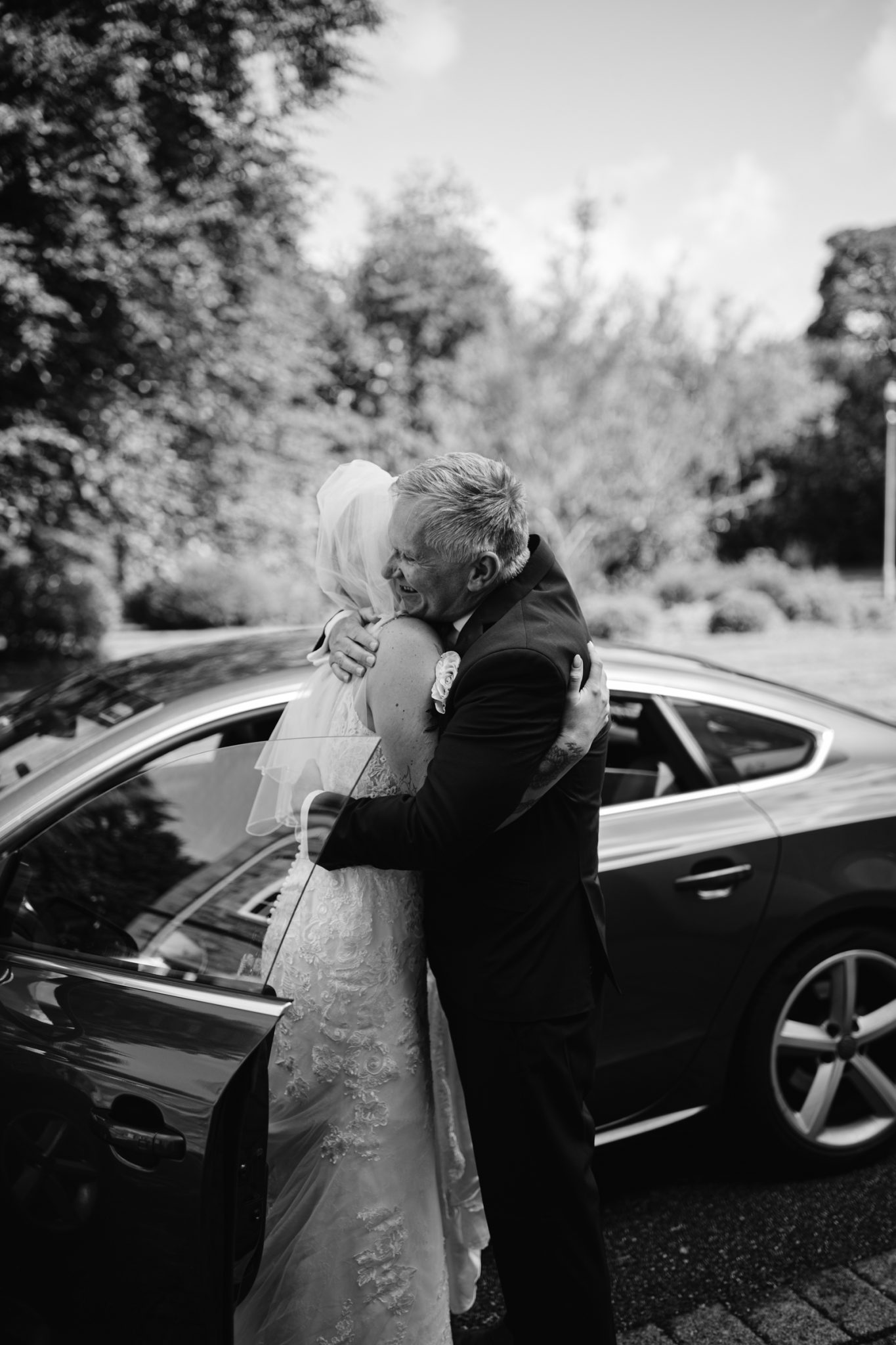 A bride in a wedding dress and veil hugs an older man in a suit beside a parked car on a sunny day.
