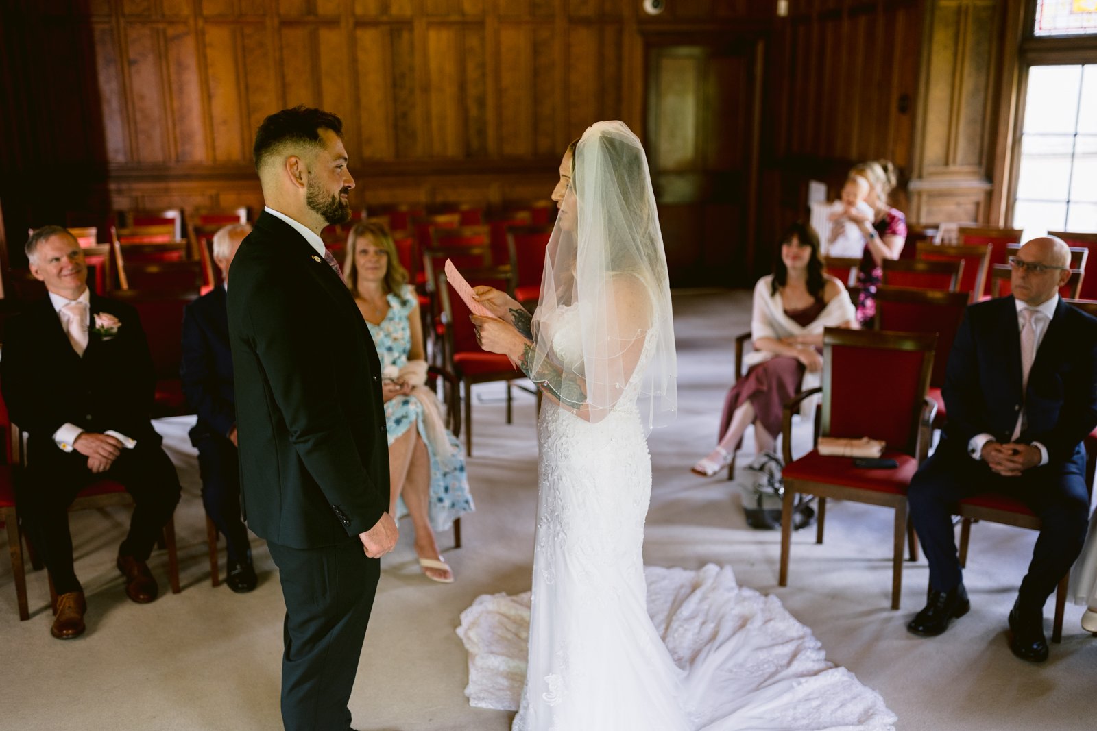 A bride in a white gown and veil reads from a card to the groom during a wedding ceremony in a wood-paneled room with seated guests watching.