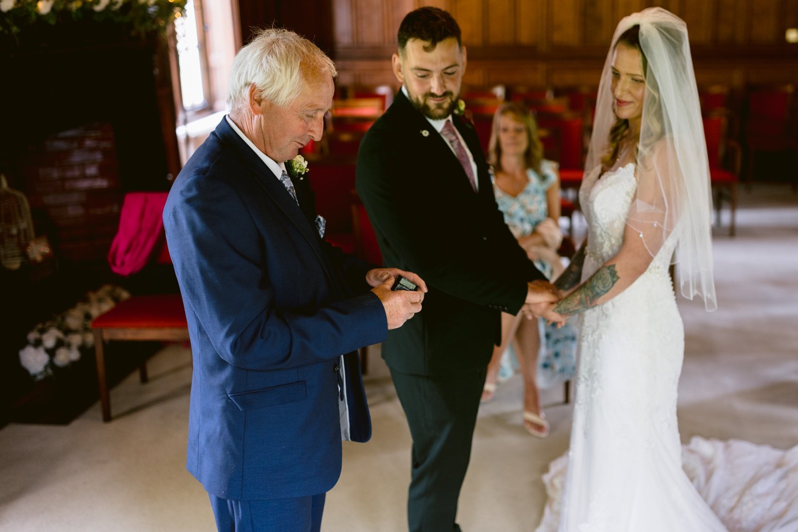 An older man in a blue suit presents a ring to a bride and groom standing together during a wedding ceremony in an indoor setting.