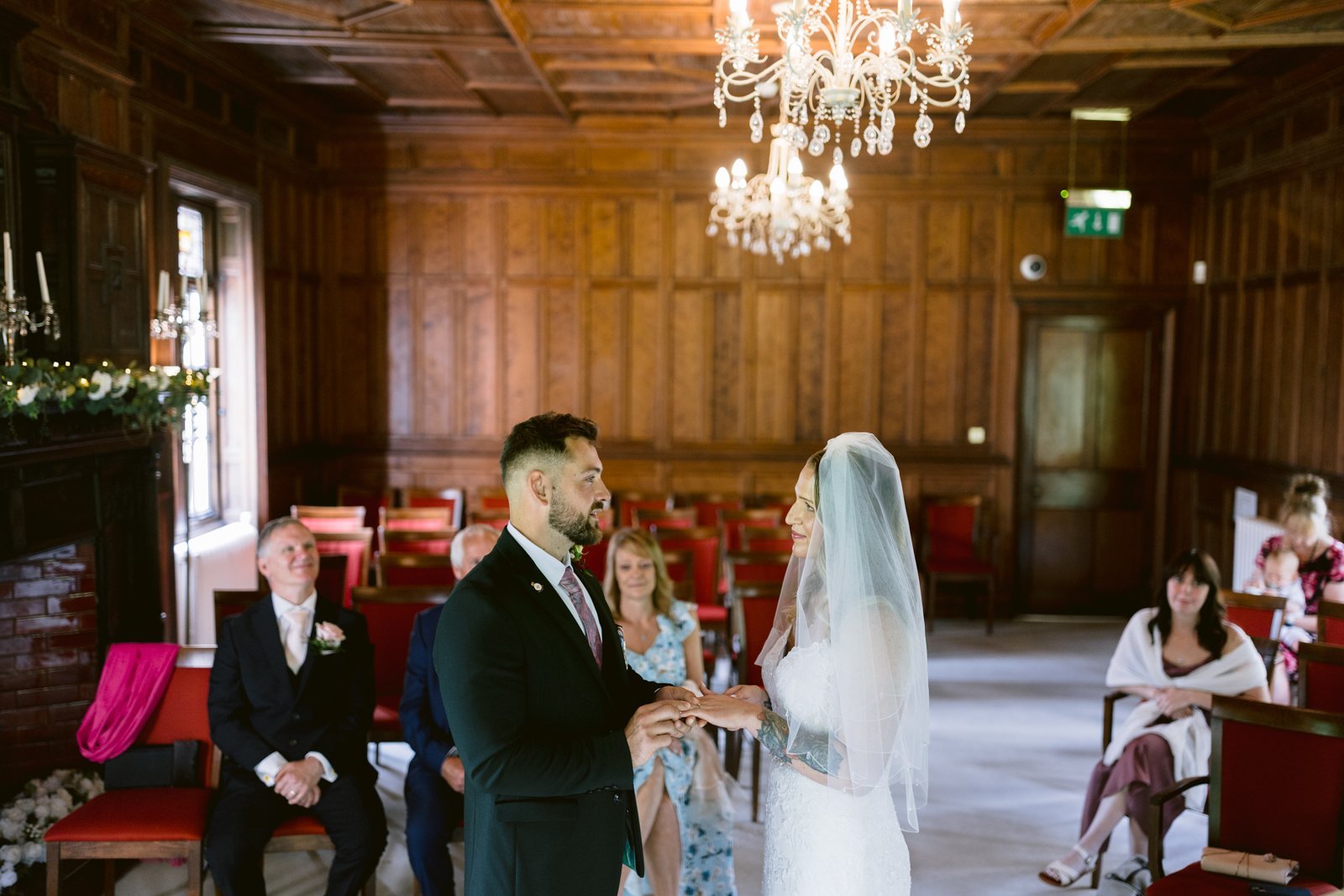 A bride and groom stand facing each other exchanging rings in a wood-paneled room with guests seated behind them.