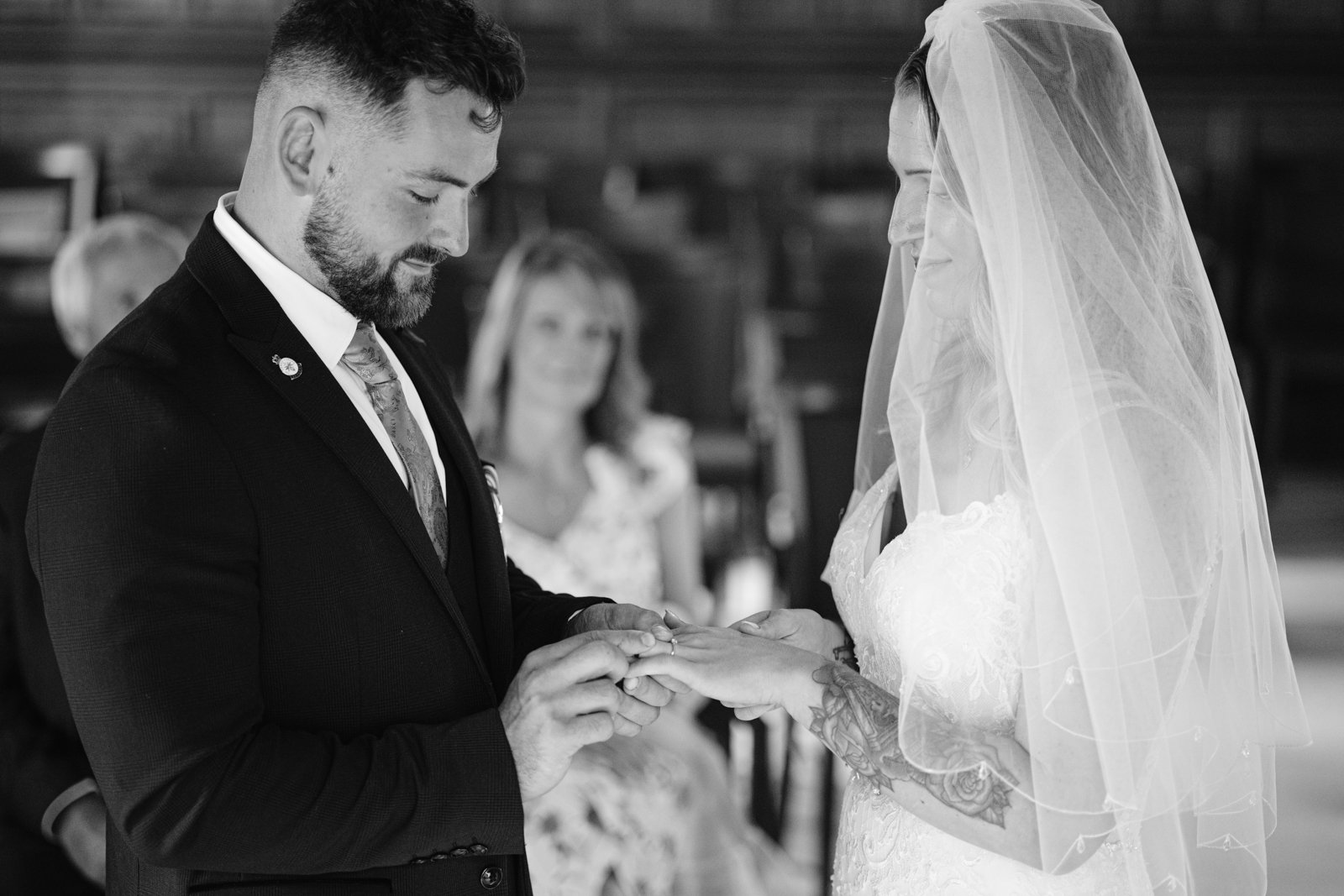 A groom places a ring on the bride's finger during a wedding ceremony; both are dressed formally, and guests are seated in the background.