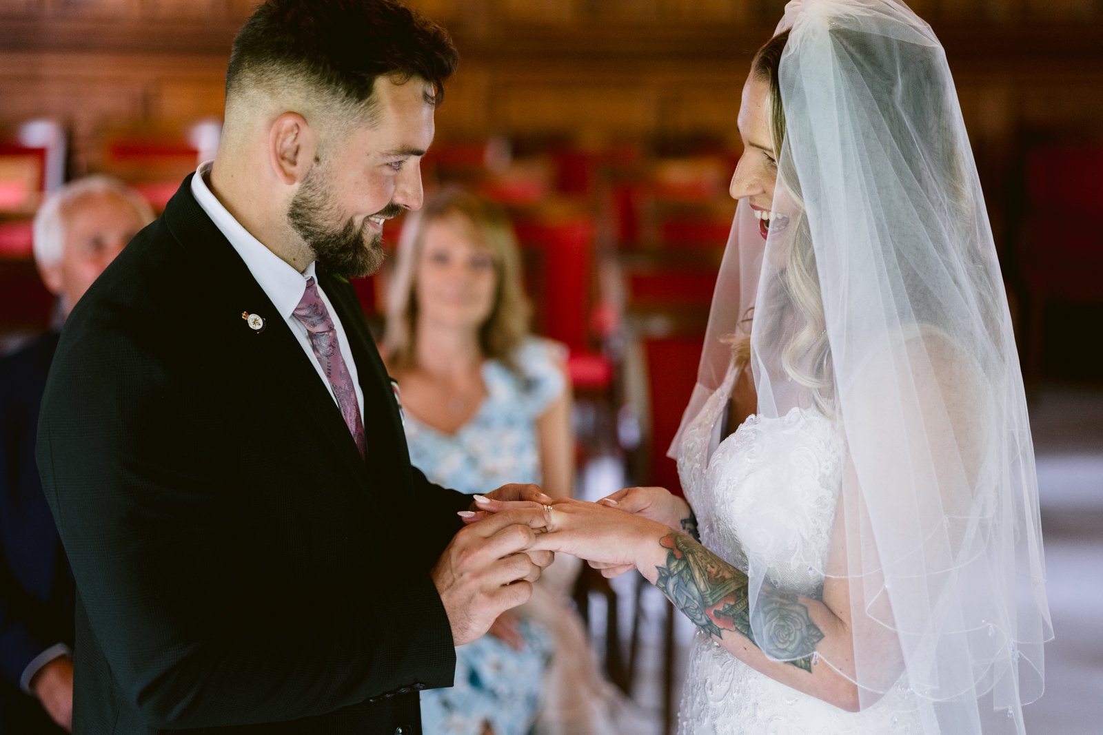 A groom places a ring on the bride's finger during a wedding ceremony, while both smile. Guests are seated in the background.