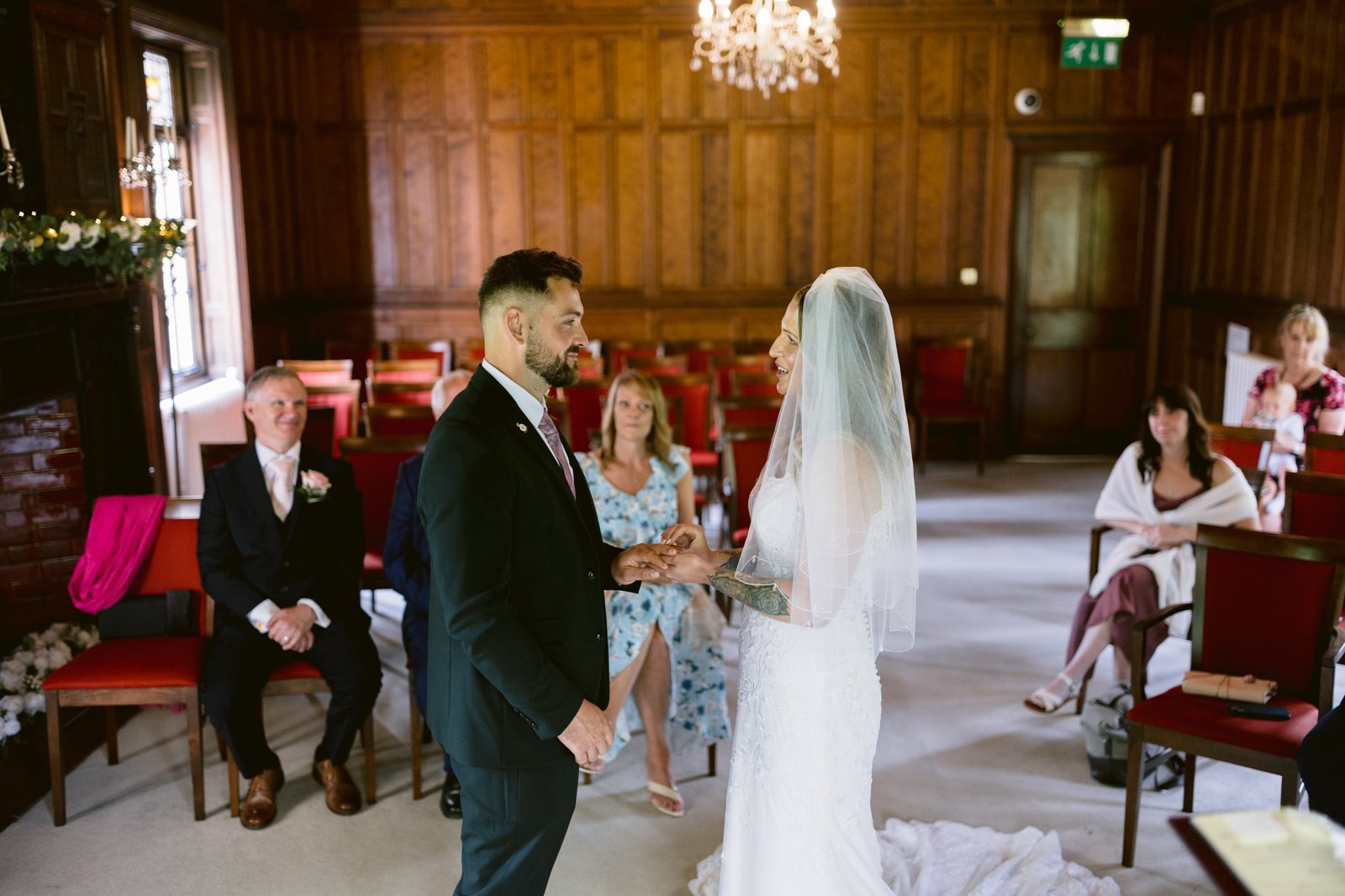 A bride and groom stand facing each other exchanging rings during a wedding ceremony in a wood-paneled room, with seated guests watching.