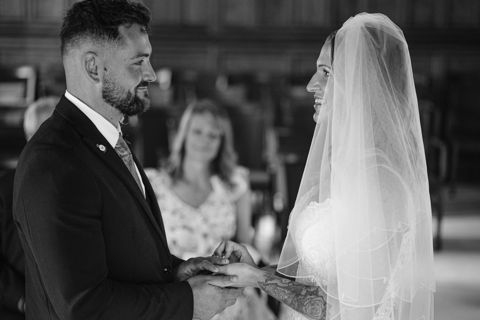 A groom places a ring on the bride’s finger during a wedding ceremony; both are smiling and facing each other, with a seated guest in the background.