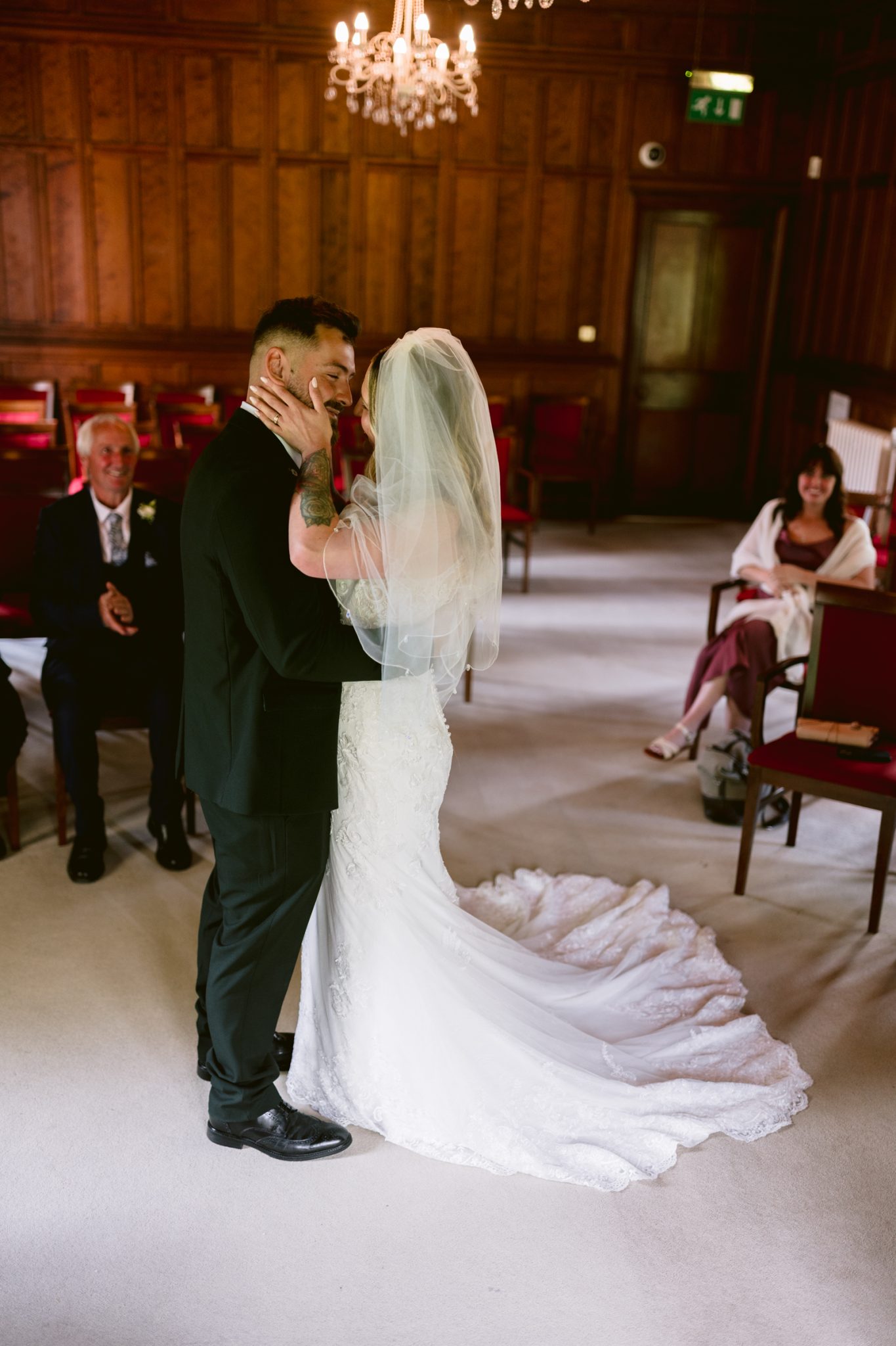 A bride and groom stand close together in a wood-paneled room, dressed in wedding attire, with a few seated guests watching.