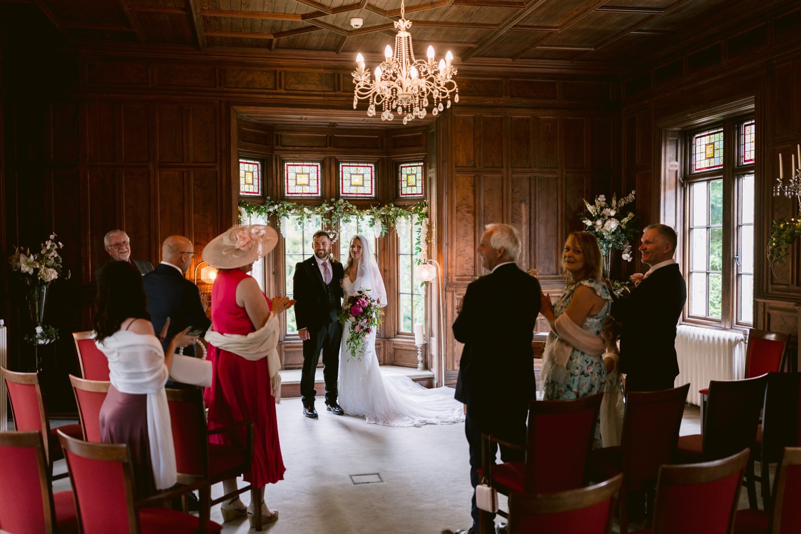 Bride and groom stand together at the front of a wood-paneled room with guests seated and standing, some applauding, during a wedding ceremony.