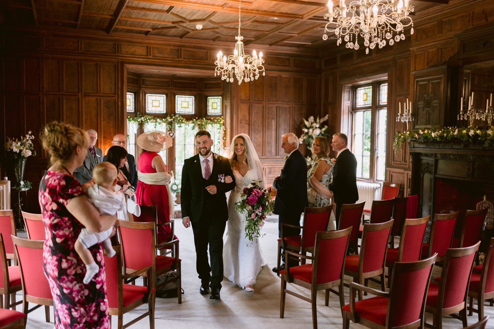 A bride walks down the aisle with a man in a suit in a wood-paneled room with red chairs, chandeliers, and guests watching.