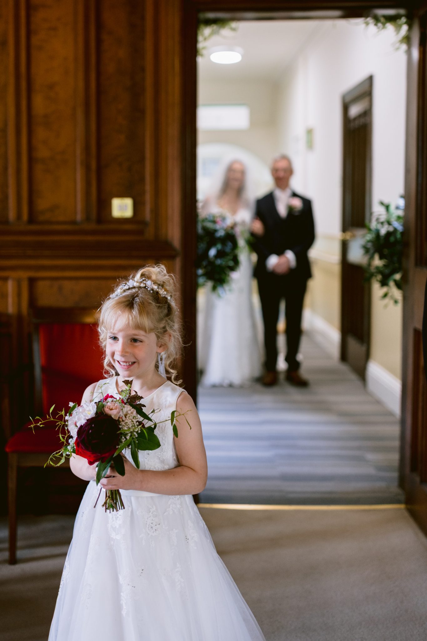 A young girl in a white dress holds a bouquet and smiles, standing in the foreground with a bride and groom blurred in the background.