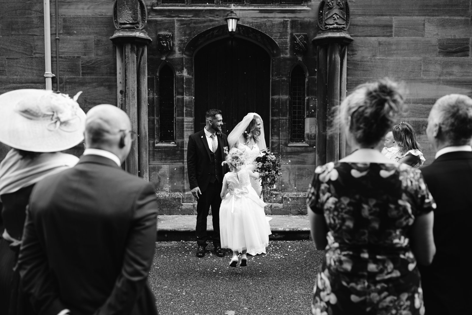 A bride, groom, and child stand outside a church door as guests in formal attire watch during a wedding ceremony.