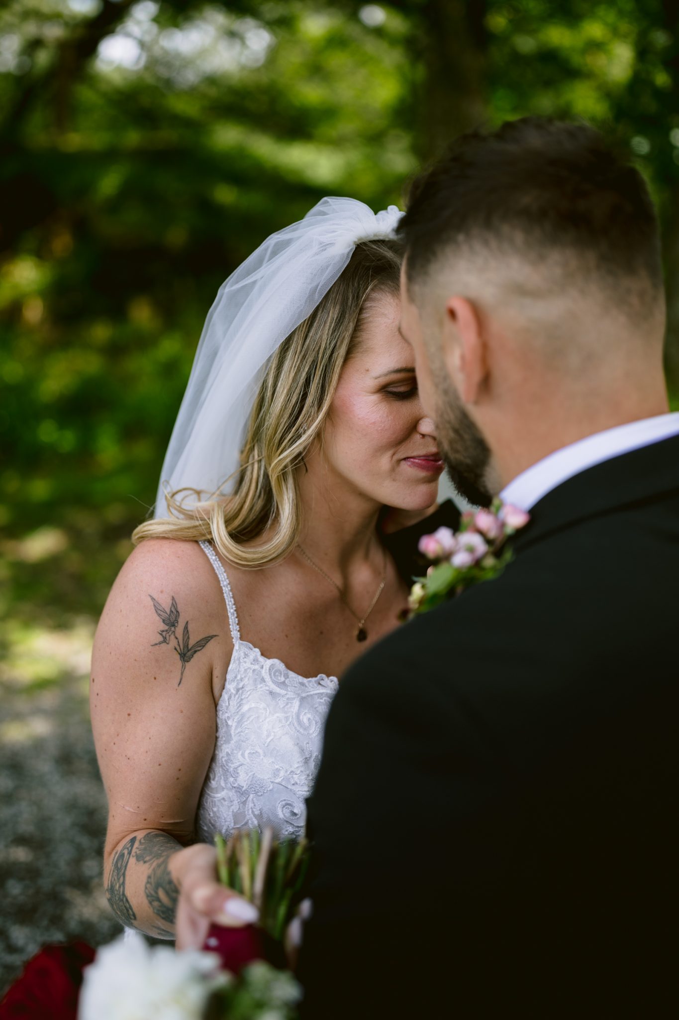 A bride in a white dress and veil stands close to a groom in a black suit, both holding small bouquets, outdoors in a natural setting.