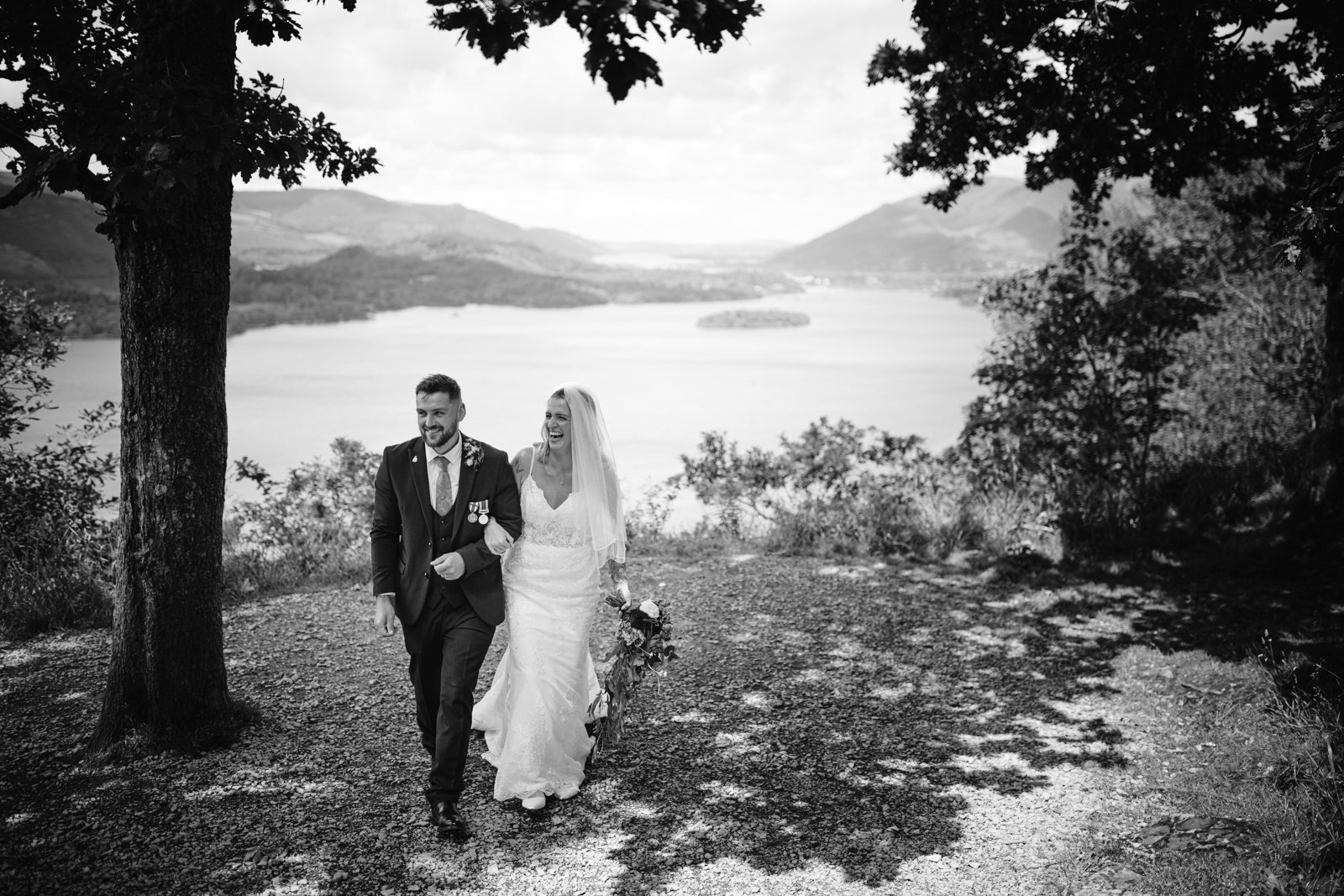 A bride and groom walk together outdoors near trees, with a scenic lake and mountains visible in the background.