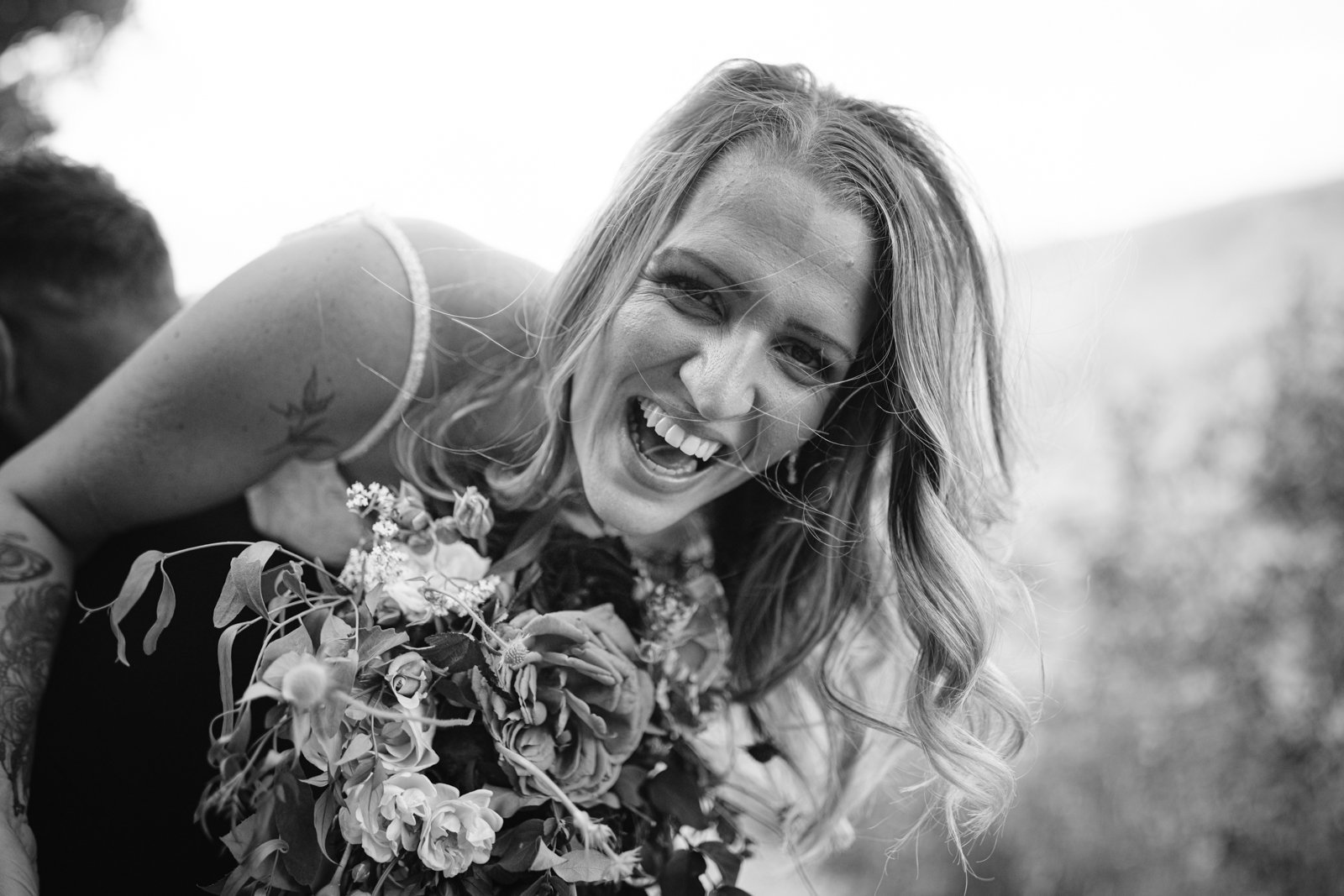 A woman holding a bouquet of flowers smiles and leans forward outdoors in a black and white photo.