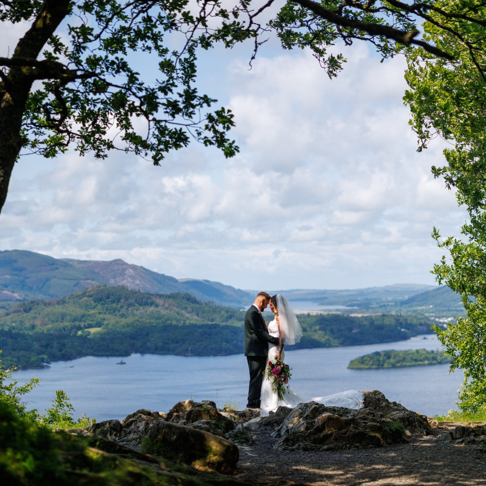 A bride and groom stand on a rocky outcrop, embracing, with a scenic view of a lake, hills, and partly cloudy sky in the background.