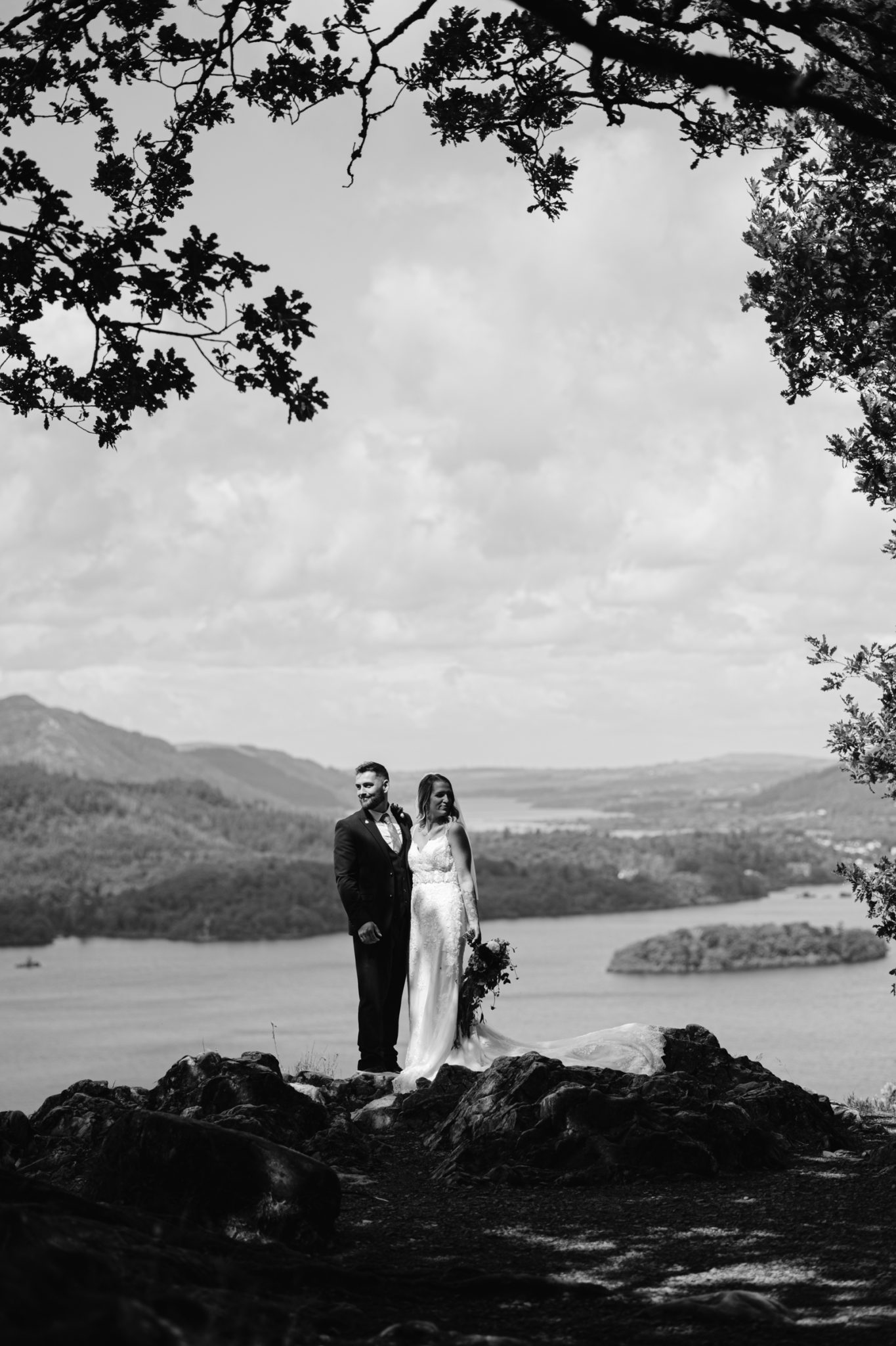 A bride and groom stand together on a rocky outcrop overlooking a scenic landscape with water and hills, framed by tree branches.