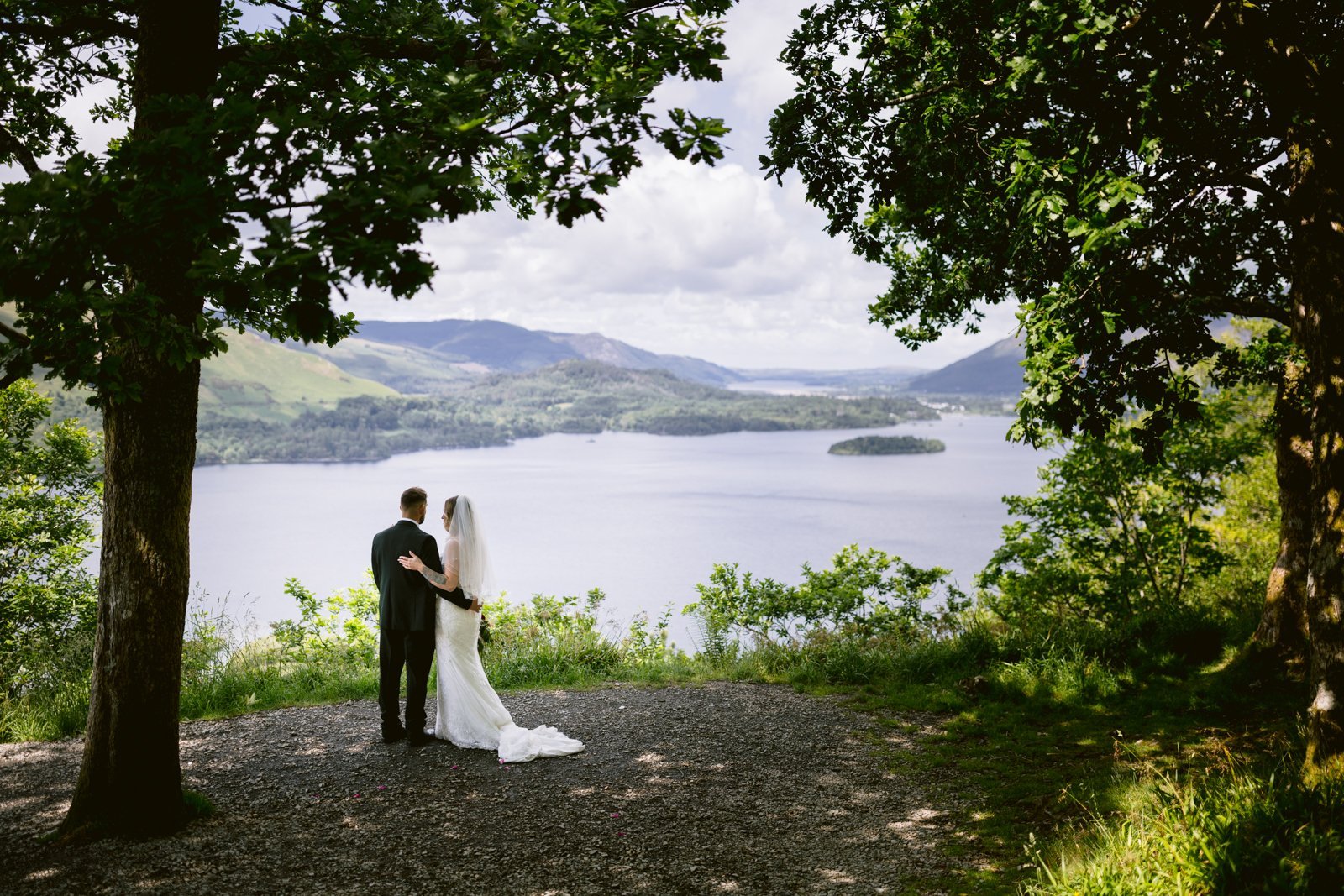 A bride and groom stand together on a hilltop, looking out over a large lake surrounded by trees and distant hills under a partly cloudy sky.