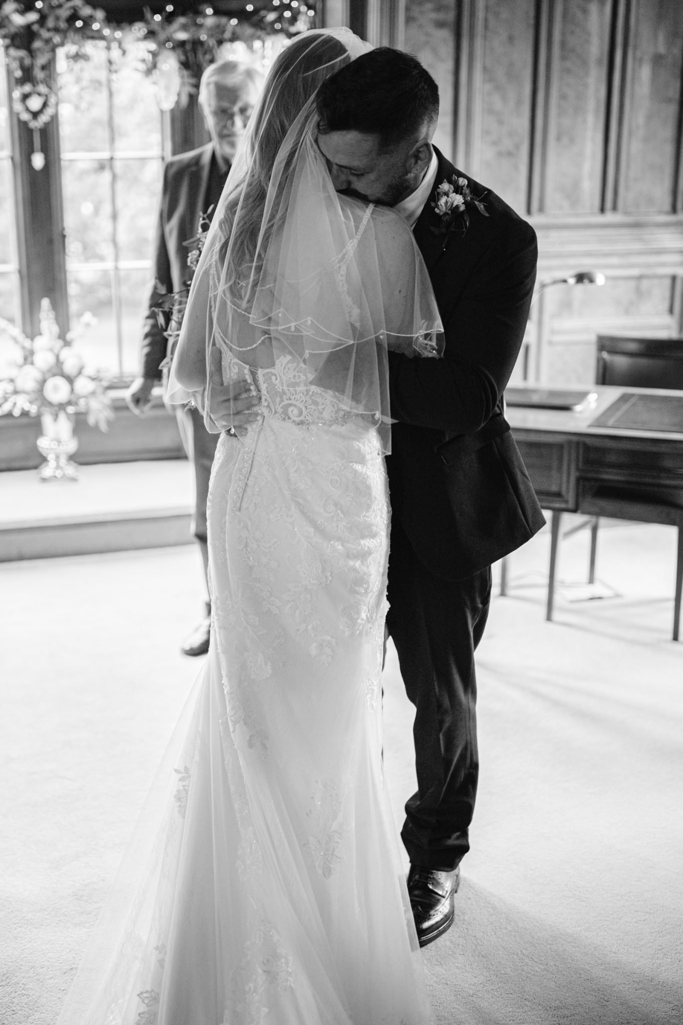A bride and groom embrace during a wedding ceremony in a room with wood paneling and large windows; an officiant stands behind them.