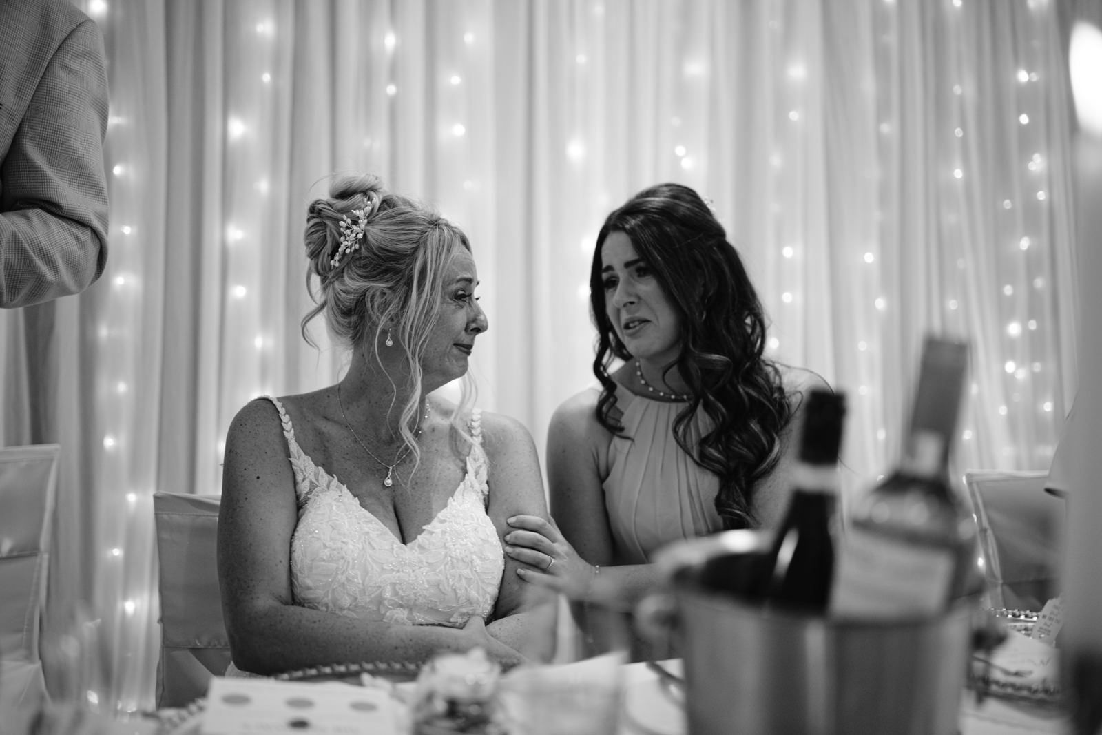 Two women sit close together at a decorated table at Armathwaite Hall, one in a wedding dress and the other comforting her, as string lights glow softly in the background of this August summer wedding.