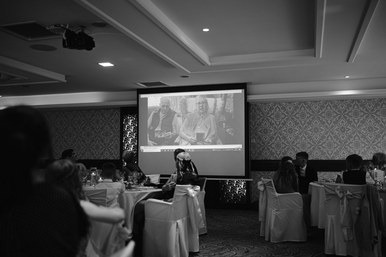 Guests seated at round tables in Armathwaite Hall’s banquet room watch a black and white video on a large screen, capturing memories from an unforgettable August Summer Wedding.