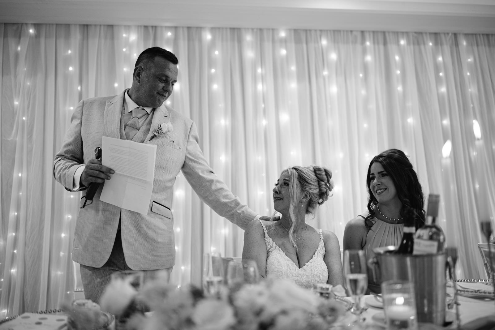 At an August Summer Wedding at Armathwaite Hall, a man in a suit gives a speech, resting his hand on the bride's shoulder; another woman sits beside them at a table with drinks, all framed by glowing string lights.