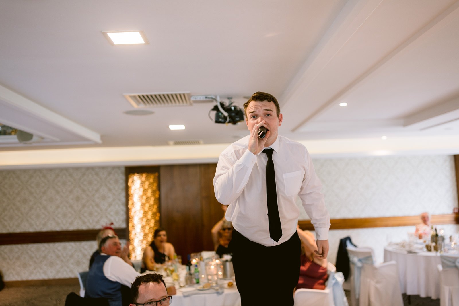 At an August summer wedding at Armathwaite Hall, a man in a white shirt and black tie stands on a chair, holding a microphone and speaking, with seated guests and decorated tables in the background.