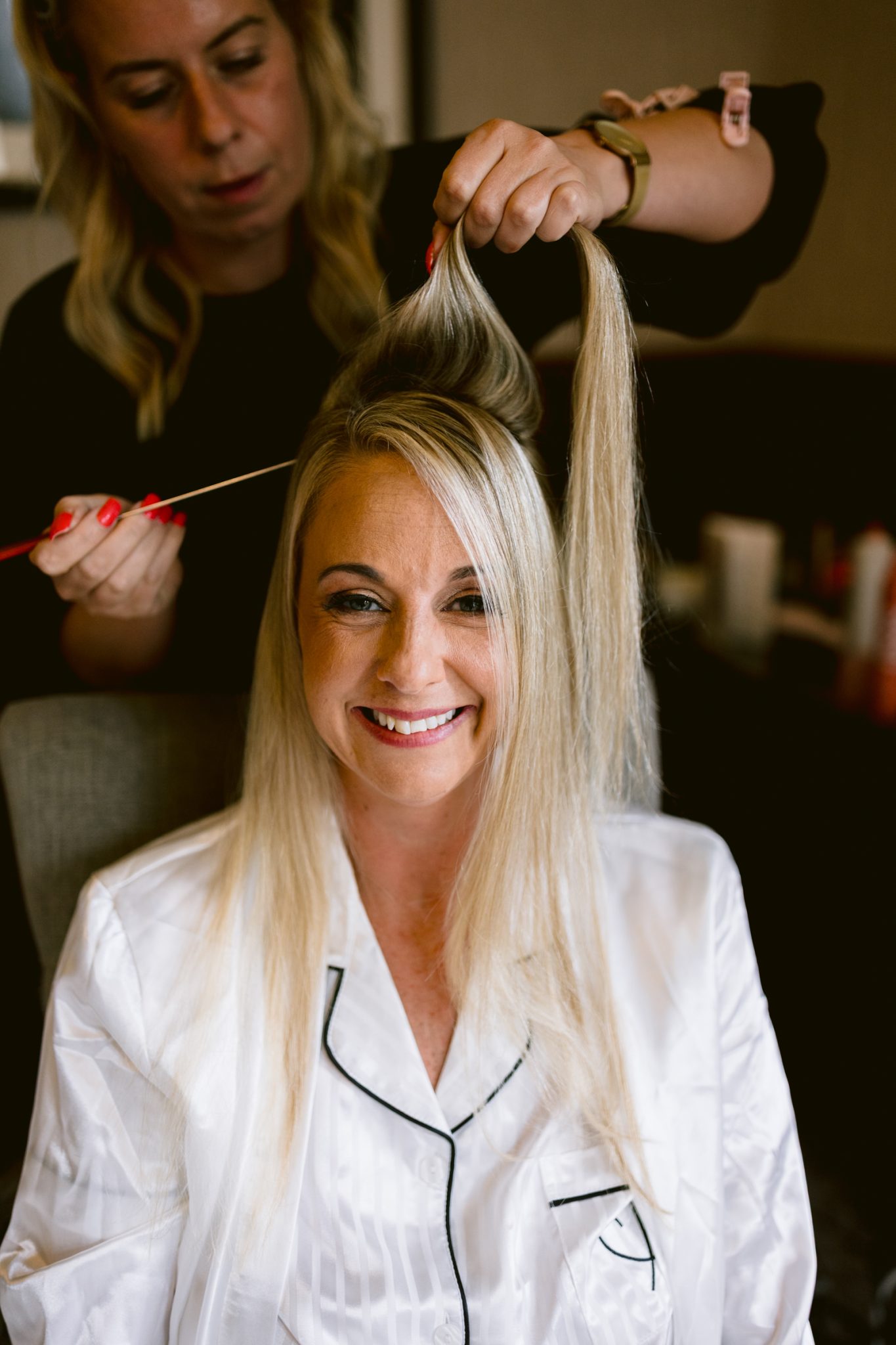 A woman in white pajamas smiles while a hairstylist lifts and sections her blonde hair with a comb, preparing her look for an August Summer Wedding at Armathwaite Hall.
