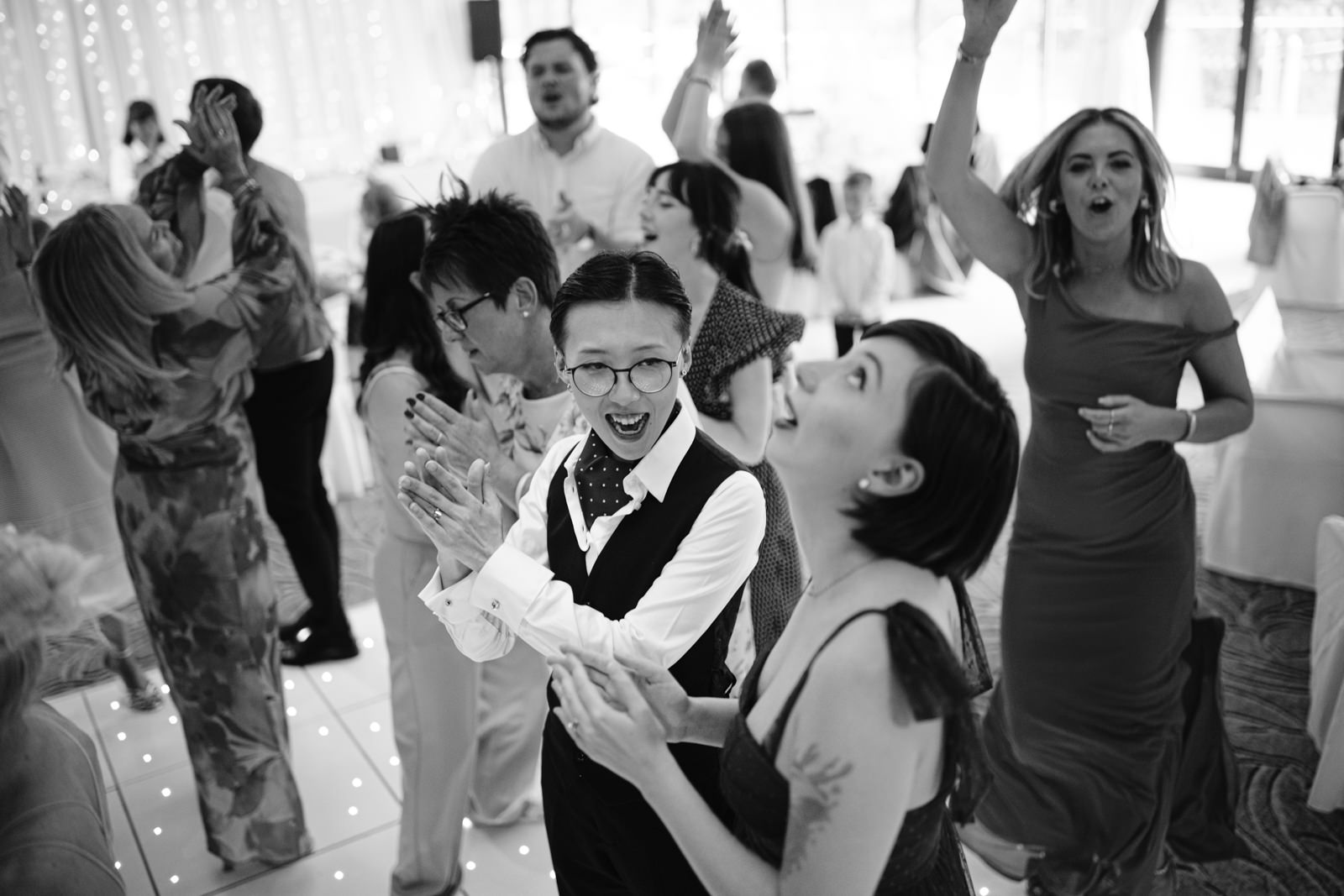 A group of people clap, sing, and dance together at an August Summer Wedding in Armathwaite Hall, with some smiling and looking up, in a lively atmosphere.