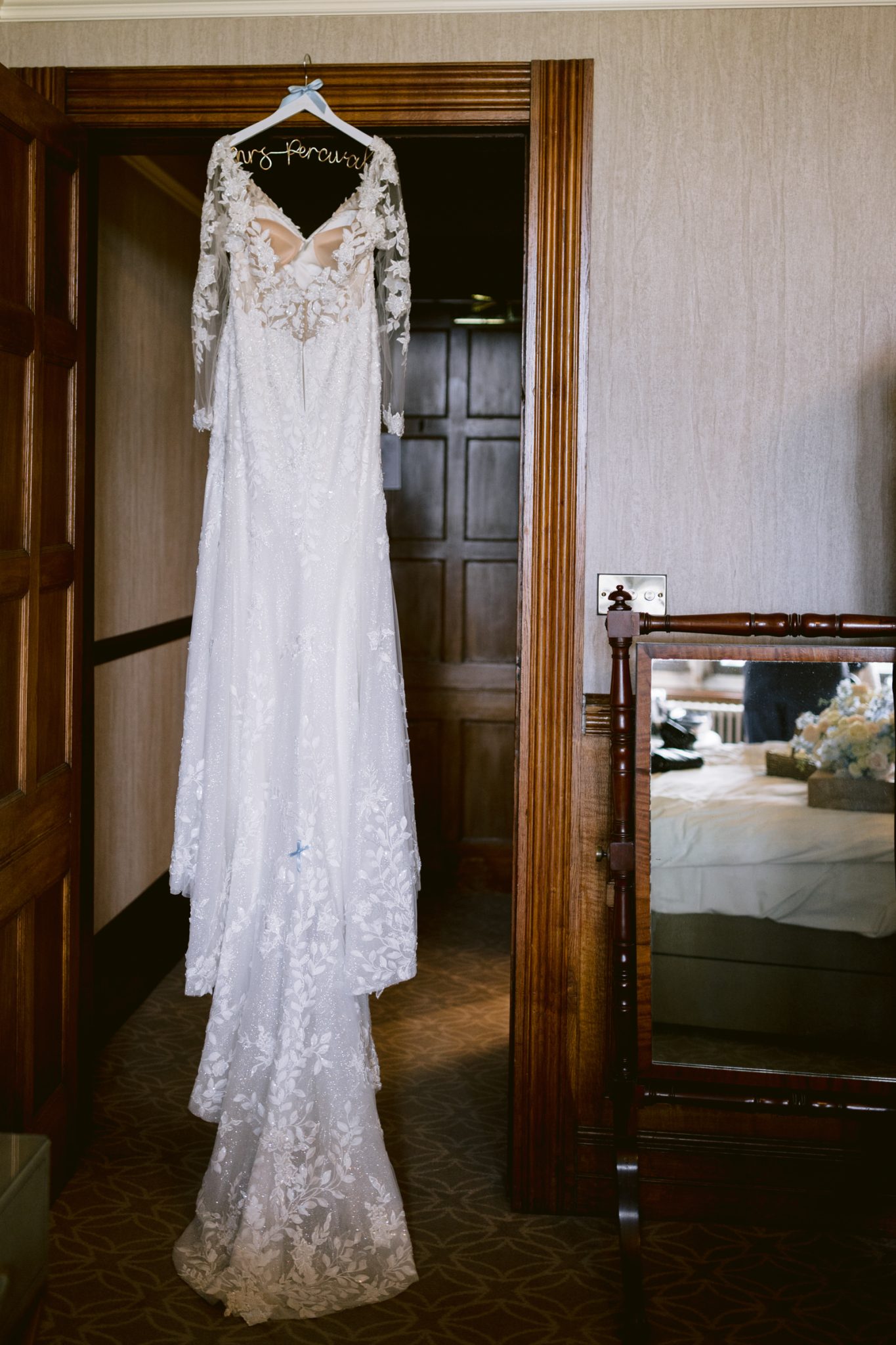 A white lace wedding dress with long sleeves hangs on a wooden hanger in a doorway at Armathwaite Hall, perfectly capturing the elegance of an August Summer Wedding. A mirror and bed are visible in the background.