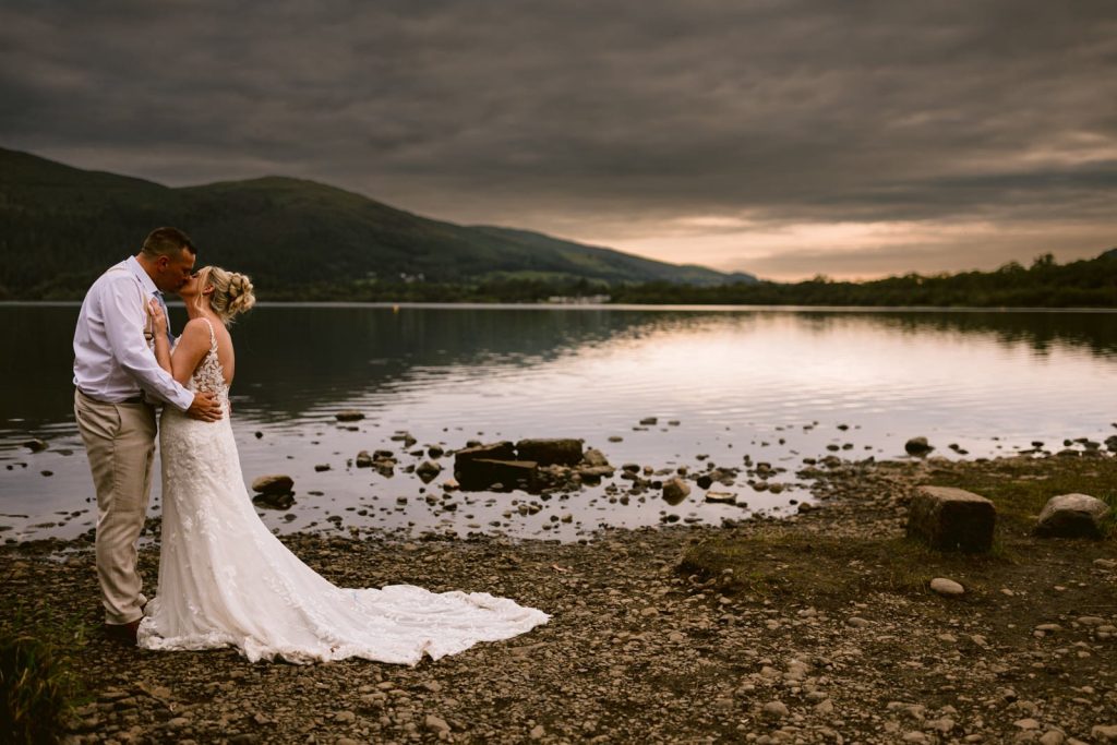 A bride and groom share a kiss on a rocky lakeshore at sunset during their August summer wedding, with mountains and a cloudy sky in the background near Armathwaite Hall.