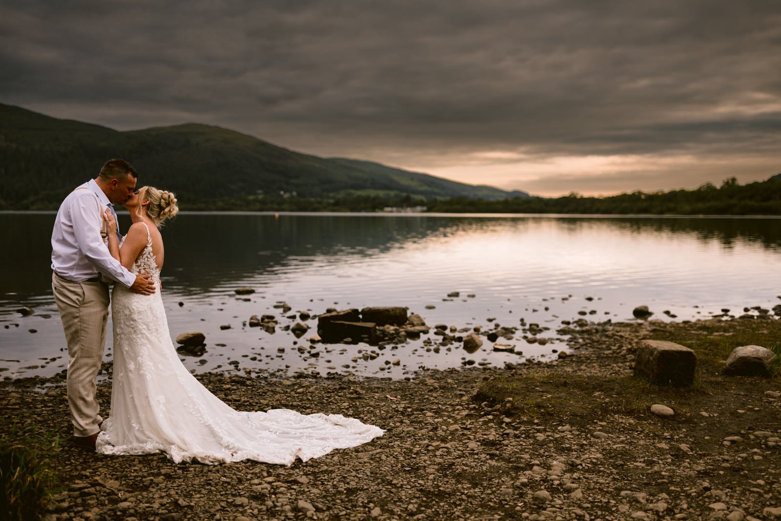A bride and groom share a kiss on a rocky lakeshore at sunset during their August summer wedding, with mountains and a cloudy sky in the background near Armathwaite Hall.