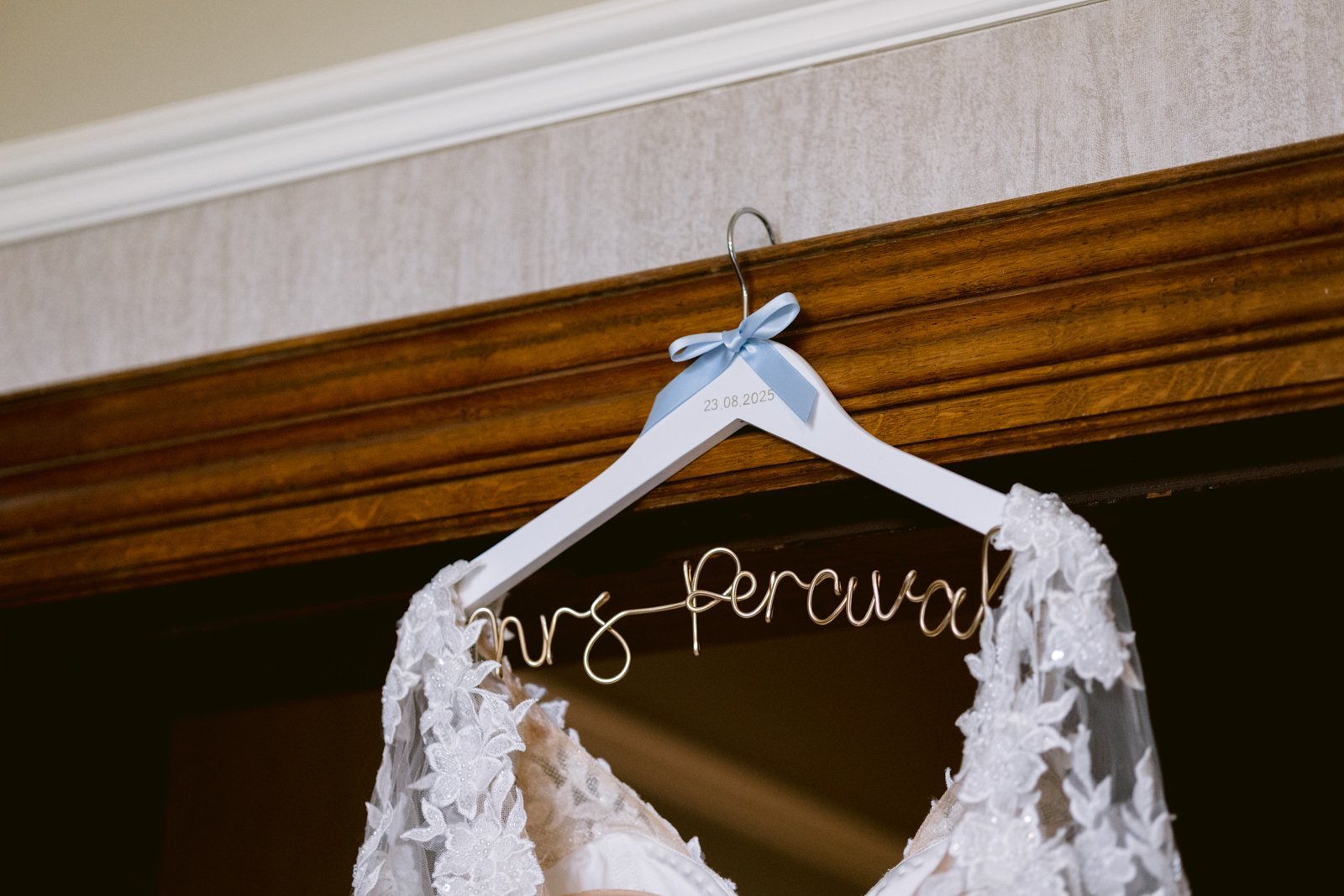 A white wedding dress with lace details hangs on a personalized white hanger with a blue ribbon, featuring a wire name and engraved date "23.08.2023"—perfect for an August Summer Wedding at Armathwaite Hall.